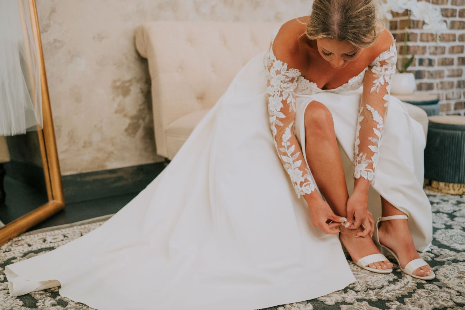 Bride in white wedding dress adjusts her shoe while sitting on vintage couch in rustic brick room.