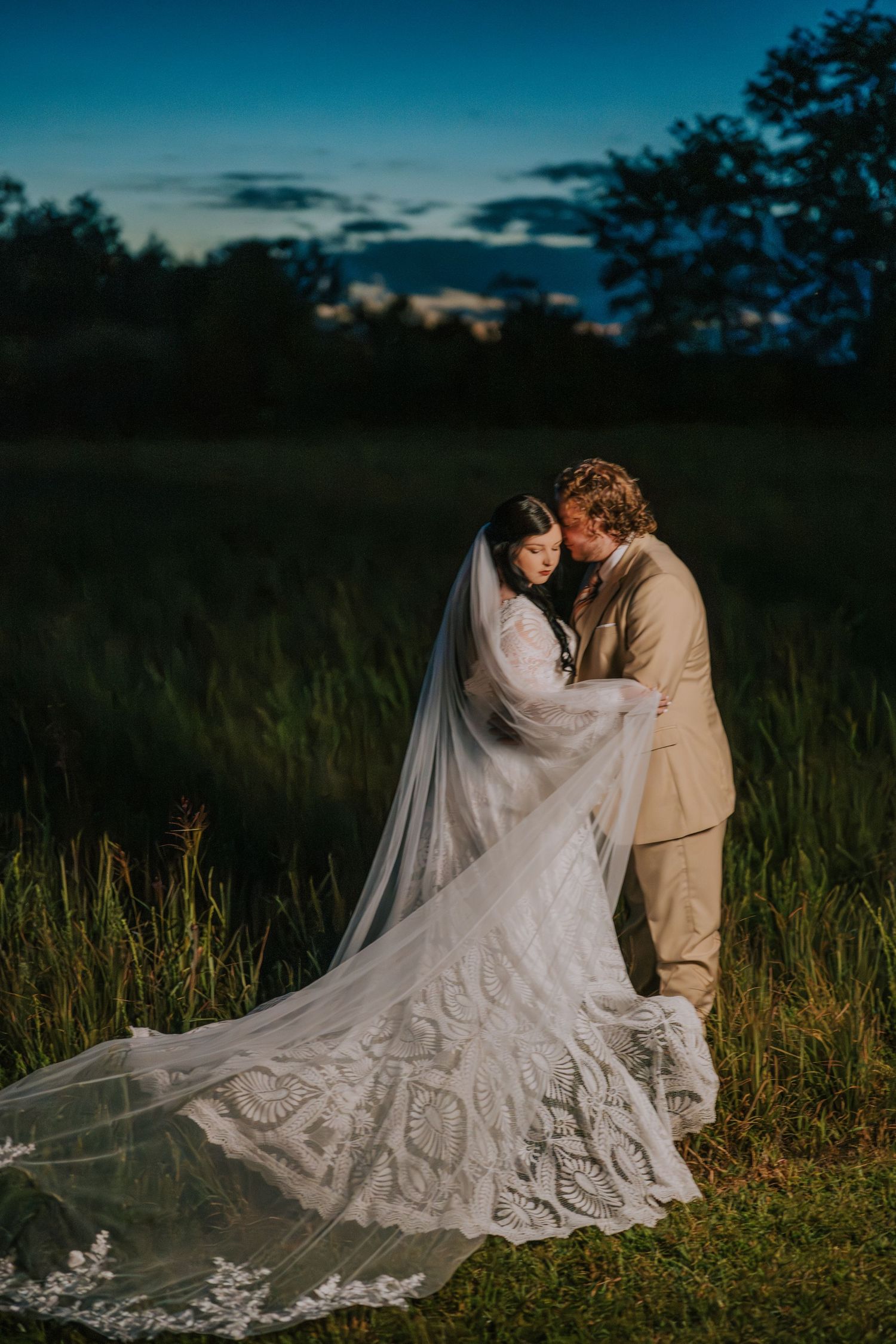 A romantic wedding couple embraces in a field of tall grass at dusk with a flowing lace veil trailing behind them.