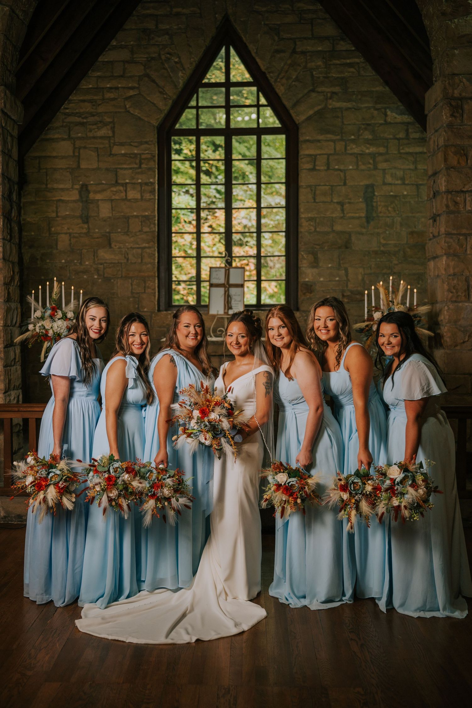 Bridesmaids in light blue dresses pose with the bride in front of a gothic stone church window.