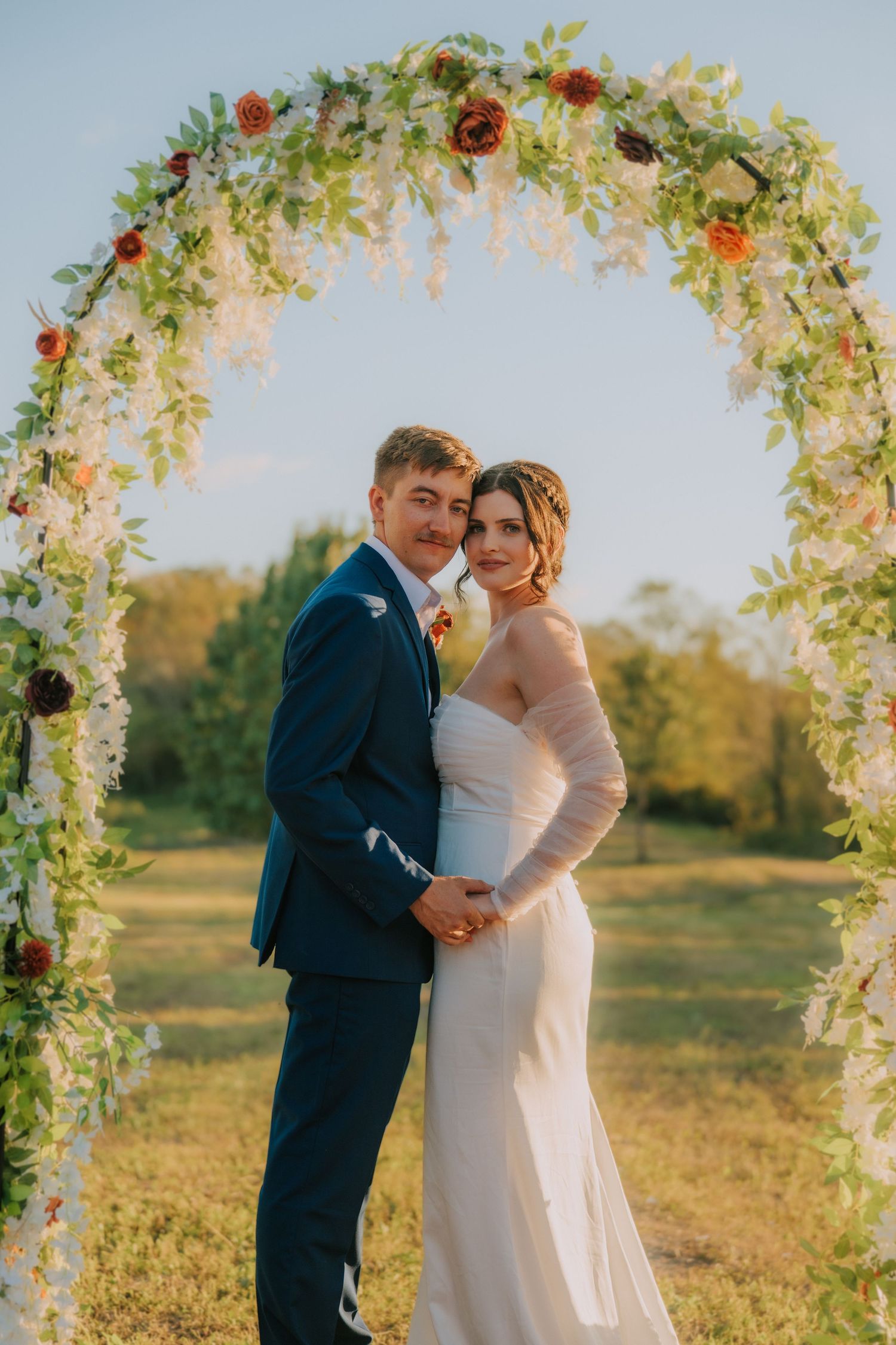 Wedding ceremony under a floral arch decorated with red roses and greenery at sunset.