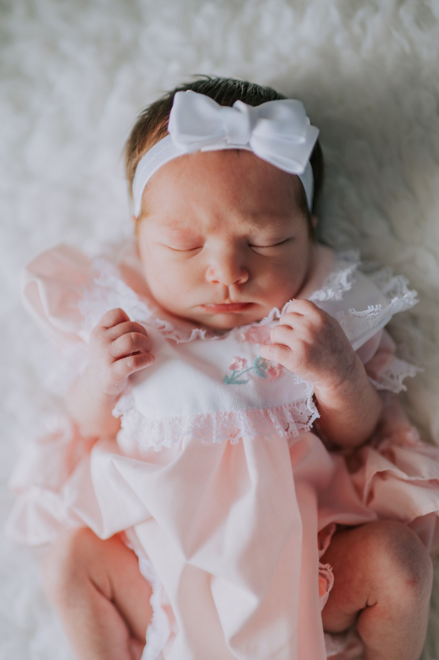 Newborn baby wearing delicate white lace dress and bow headband on soft white backdrop.