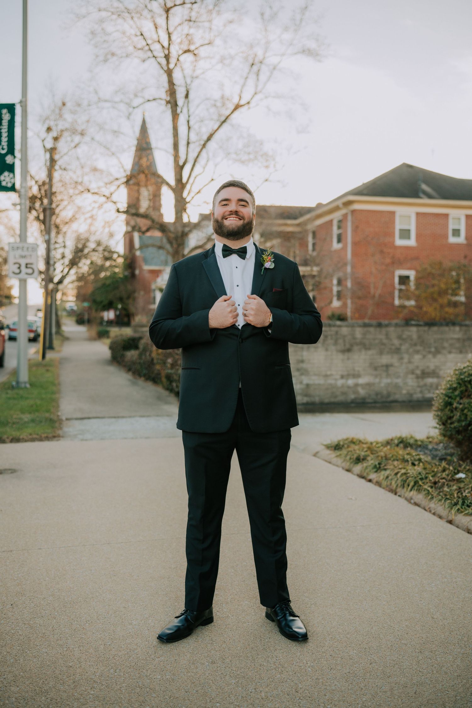 A groom in a classic black tuxedo and bowtie stands confidently on a path with a church spire visible in the background.