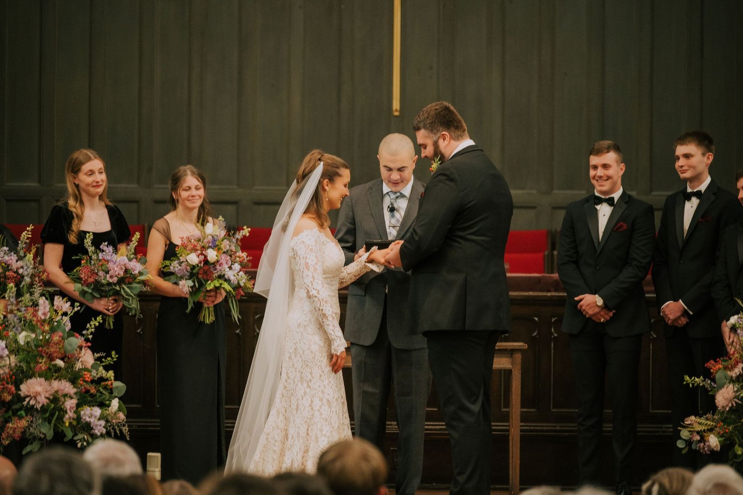 A wedding ceremony at a church altar with bridesmaids in dark dresses and pink floral arrangements.