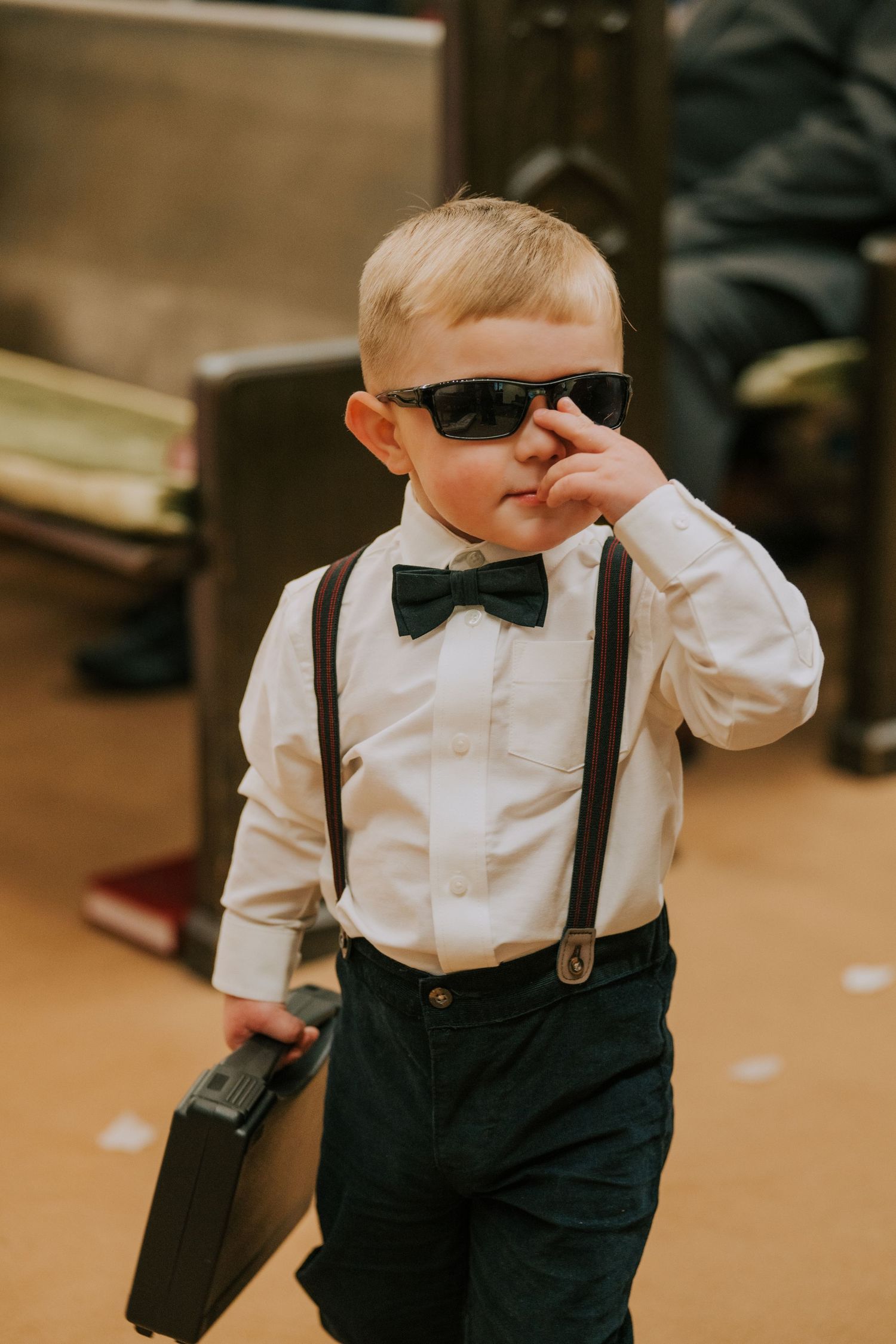 Small ring bearer in suspenders and bow tie wearing sunglasses while walking down church aisle during ceremony.
