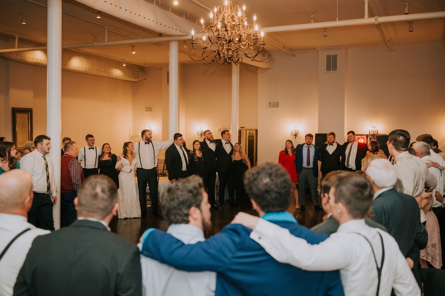 Large group of wedding guests gathered under chandelier for celebration dance.