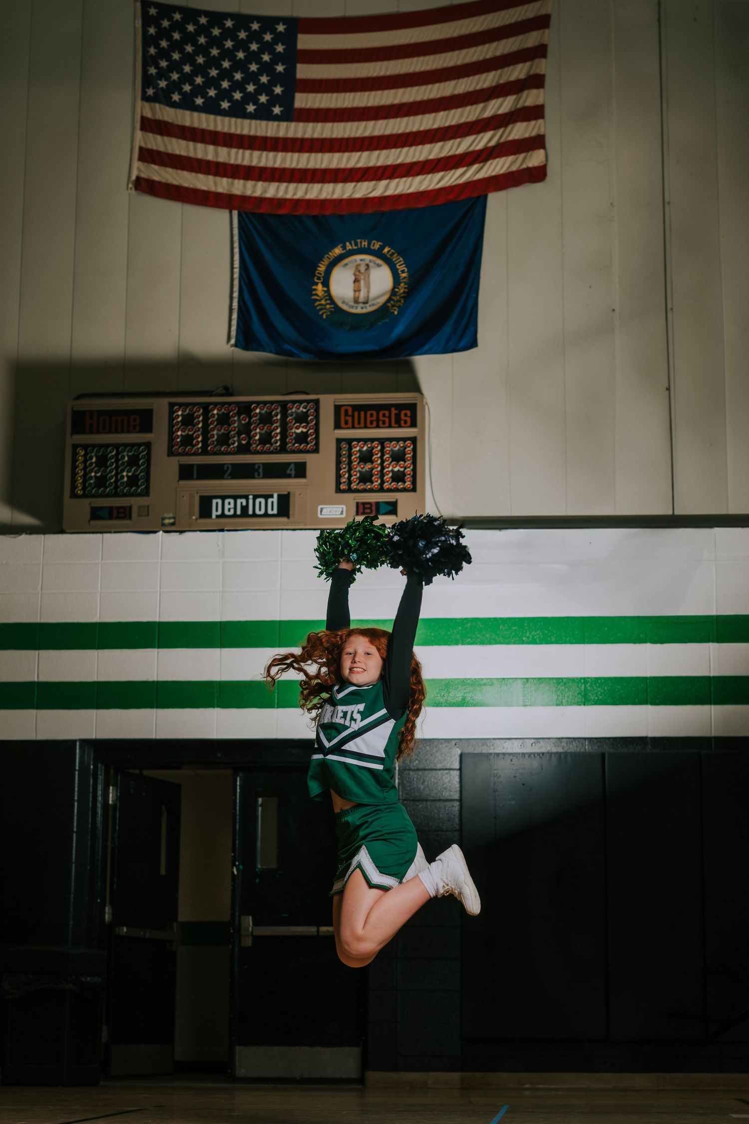 Lacrosse player in green and white uniform poses against wall with American flags and school banners displayed above.