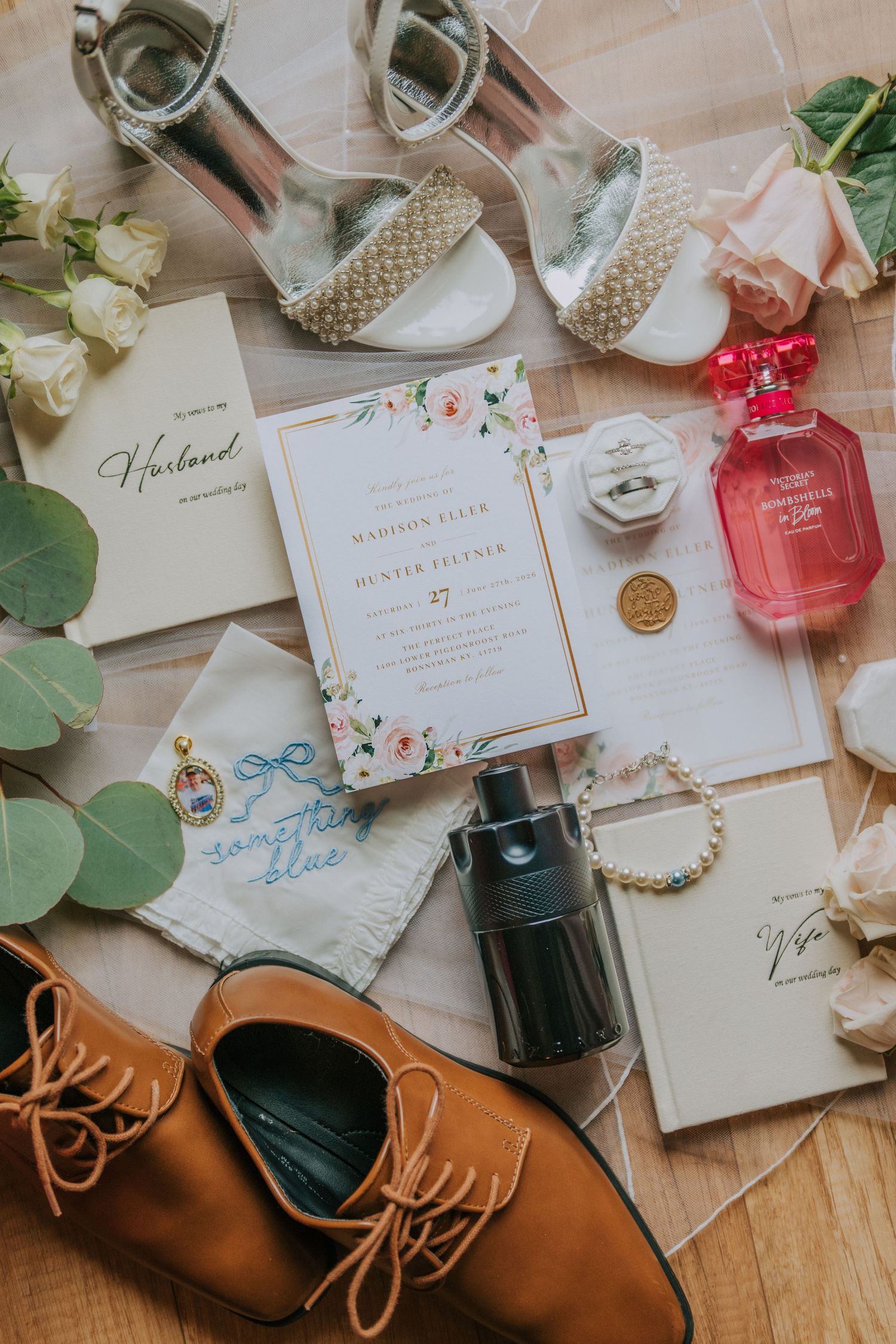 Elegant wedding flat lay with floral invitations, pearl heels, perfume, rings, and bridal accessories on wood floor.
