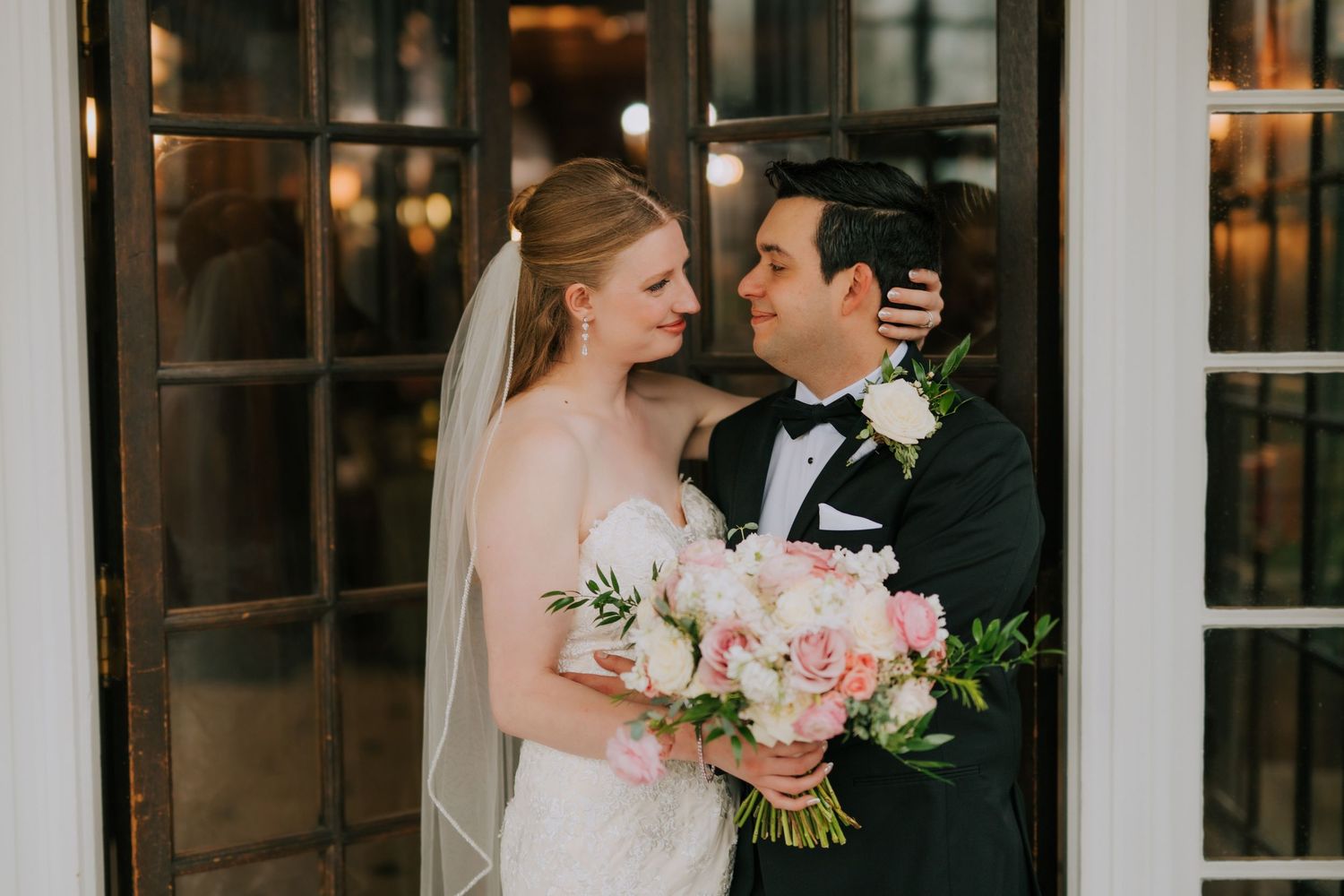 Bride and groom share a romantic moment outdoors, bride holding pink and white bouquet, groom in black tuxedo.