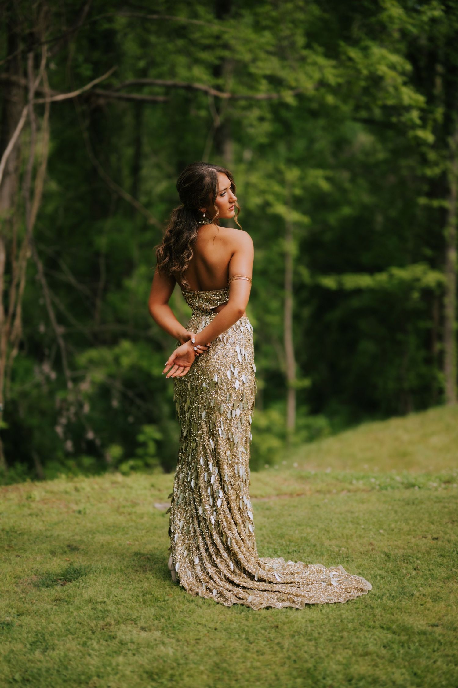 Woman in gold sequin gown poses outdoors on green grass with lush forest backdrop.