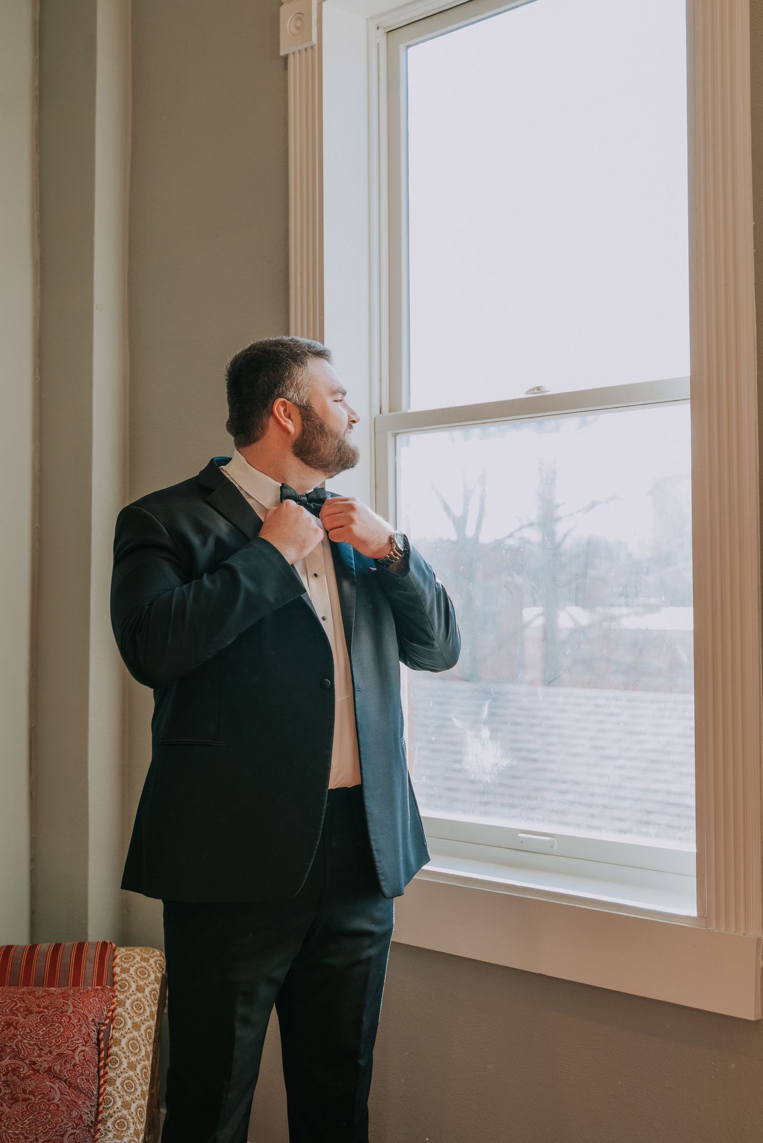 A person in a black suit adjusts their tie while standing near a tall window with natural light streaming in.