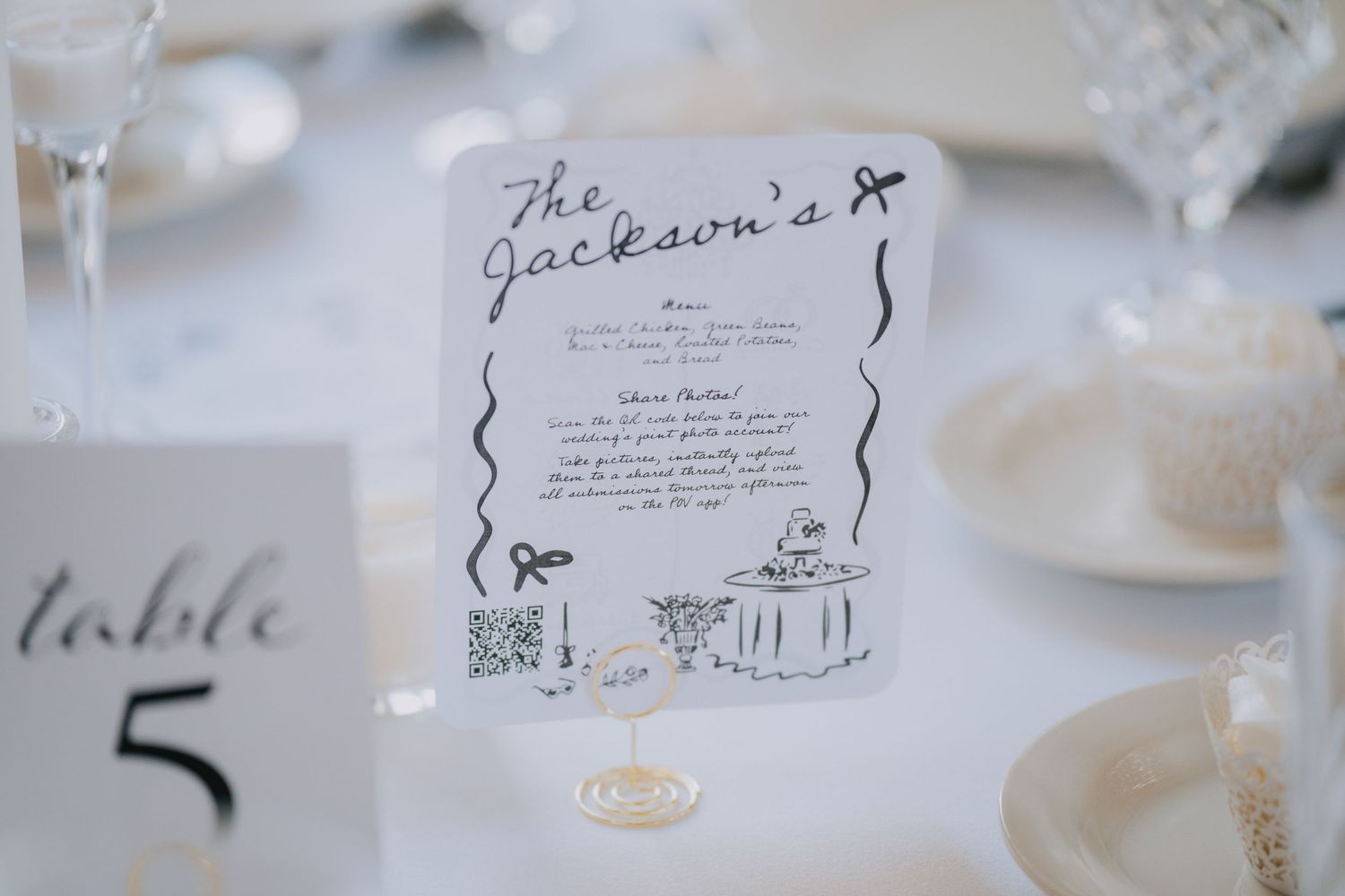 Elegant wedding menu card sits on a white tablecloth next to a table number marker at a formal place setting.