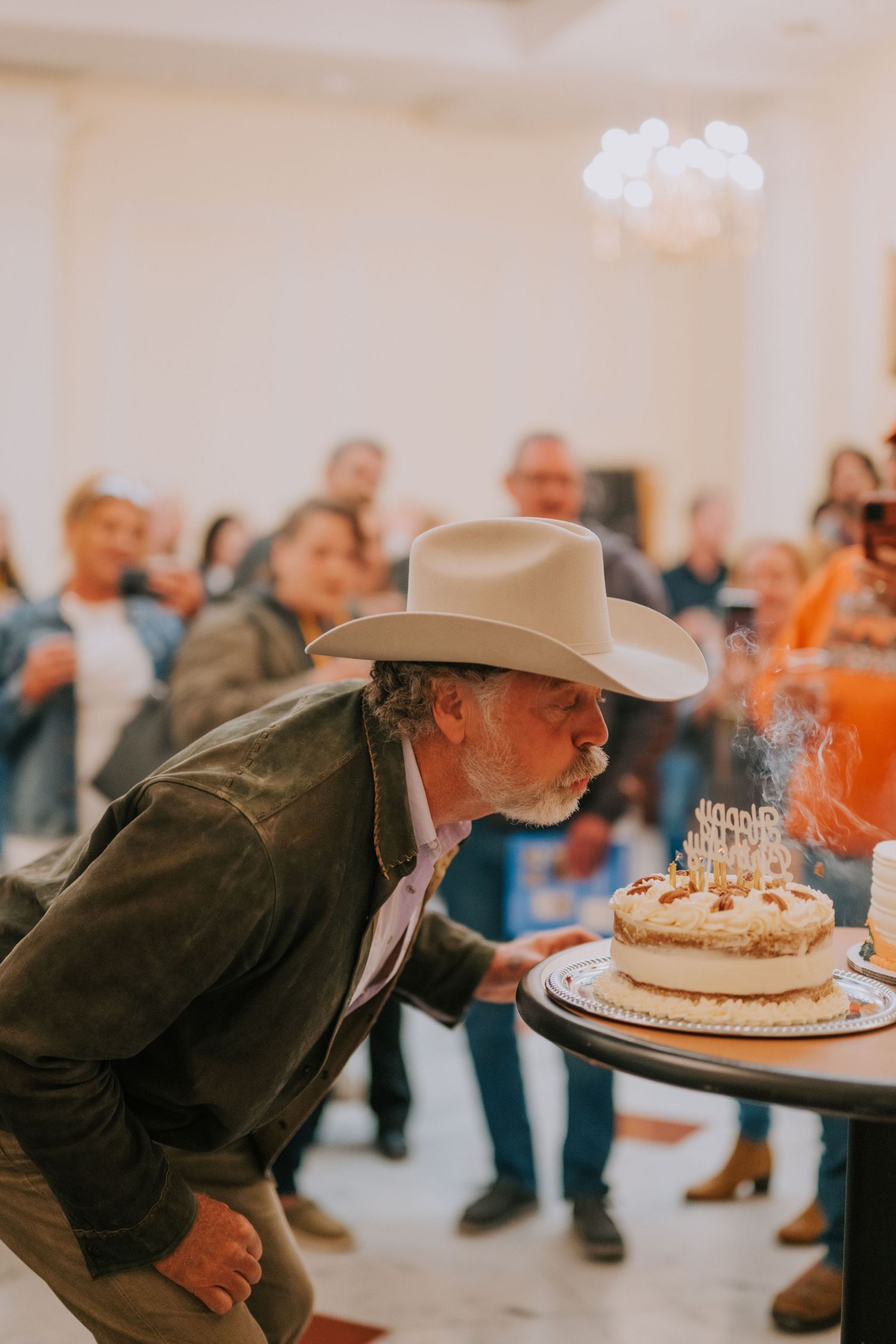 A person in a cowboy hat leans forward to blow out birthday candles on a cake while others watch in the background.