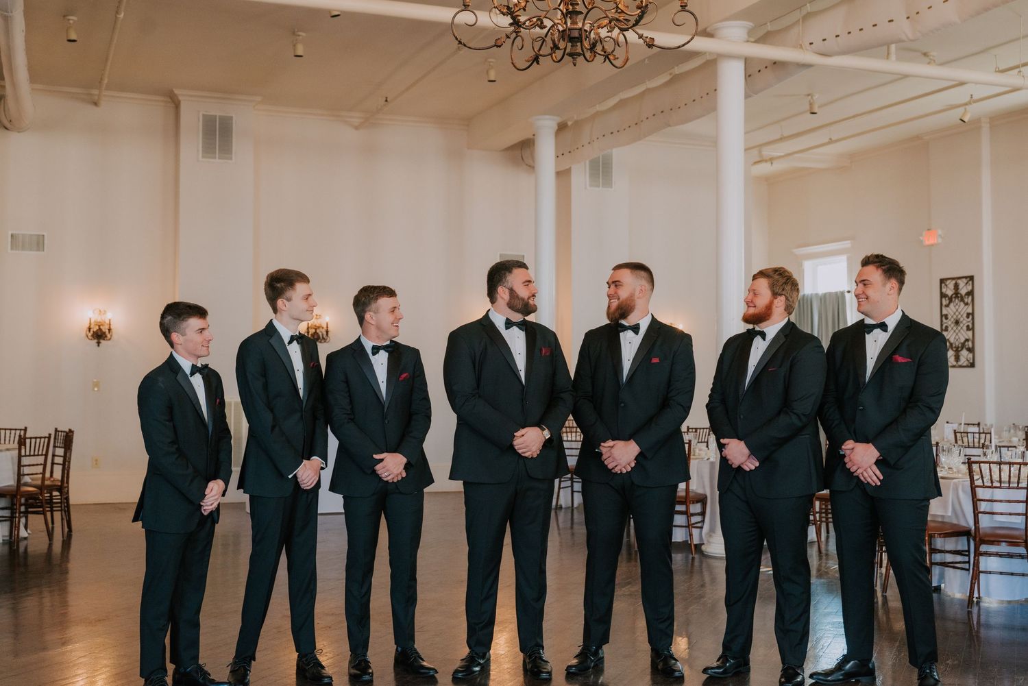 Groomsmen wearing matching black suits line up in an elegant white room with ornate architectural details.
