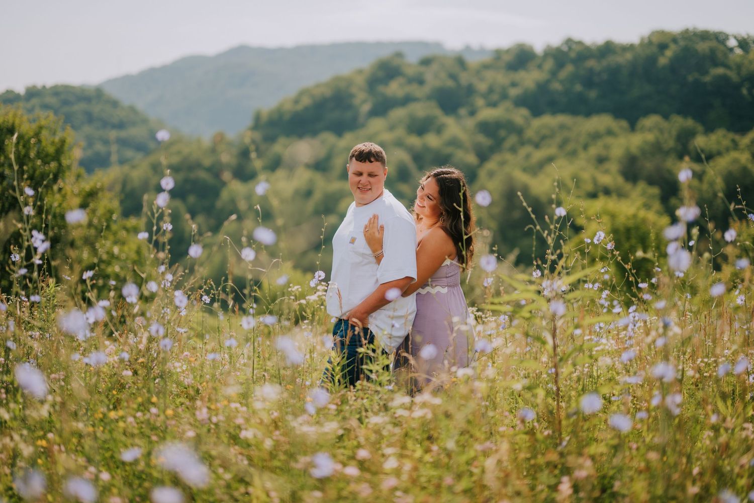A couple stands together in a field of white wildflowers with mountains in the background.