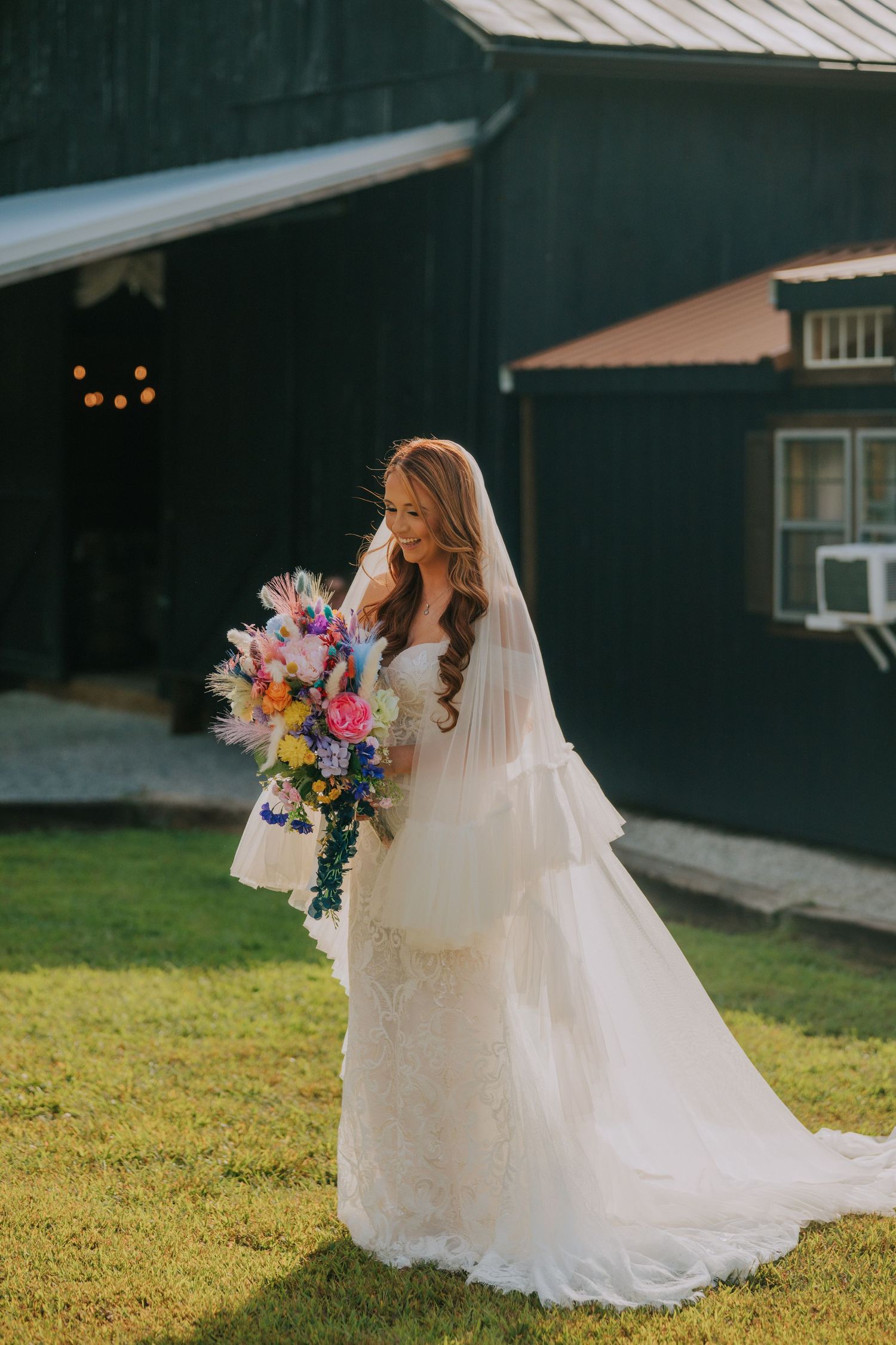 A bride in a flowing white gown holds a vibrant bouquet while standing on grass near a dark building.