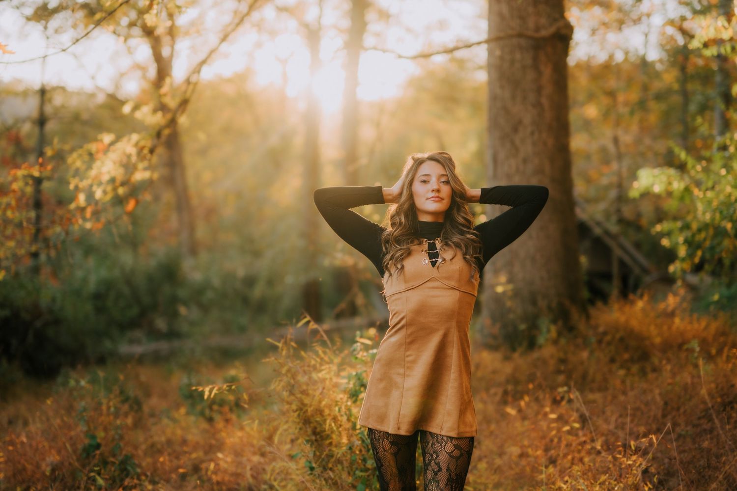 A woman in a brown dress poses in a sunlit autumn forest with golden foliage.