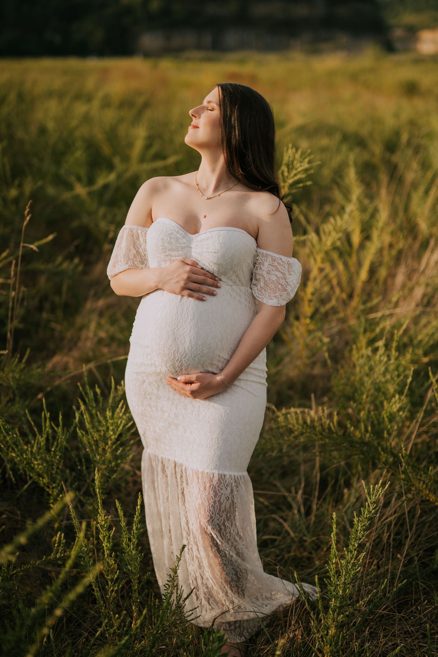 Ethereal maternity portrait in a white lace dress amid golden sunlit grass at sunset.