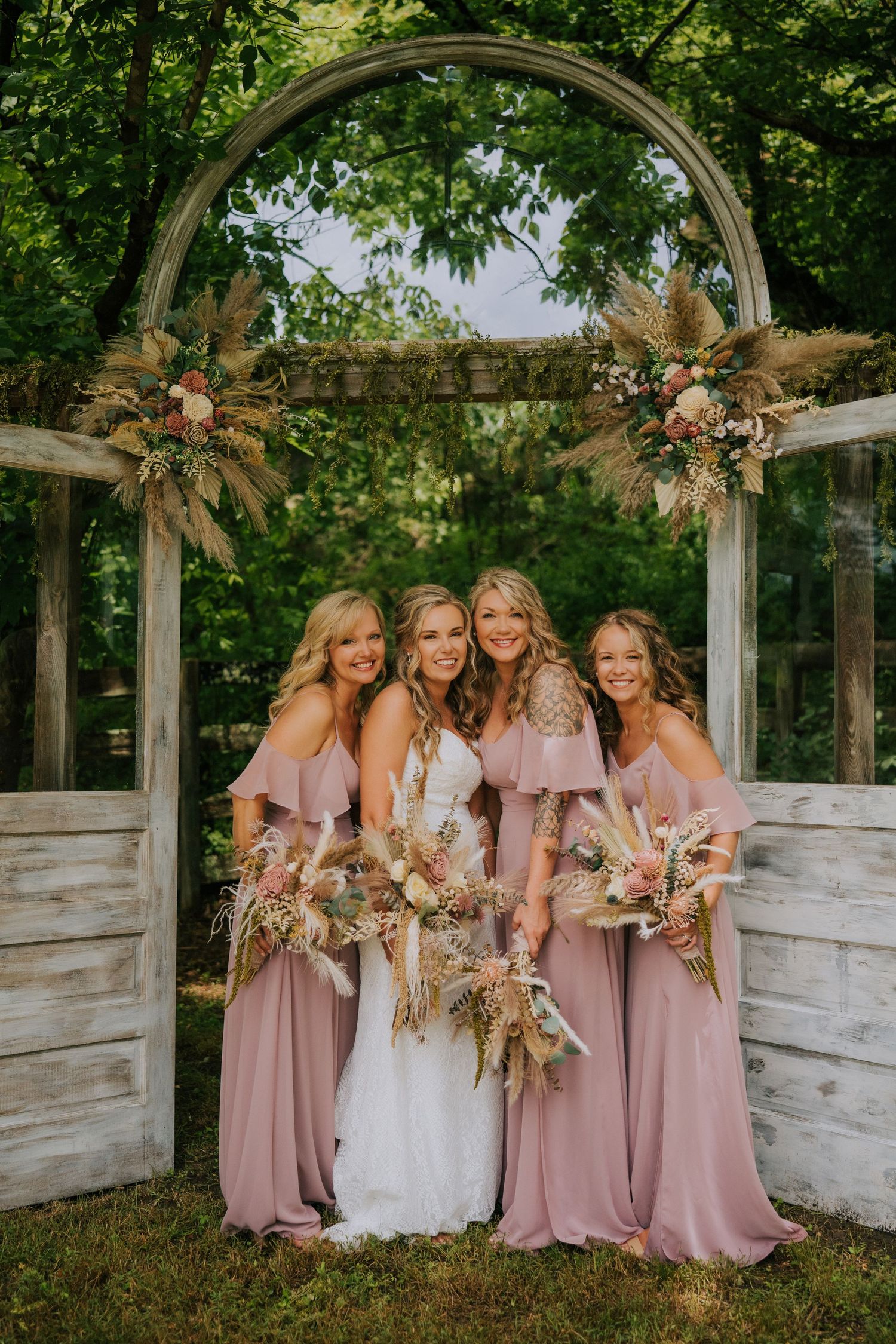 Bridesmaids in dusty rose dresses pose together under an elegant white garden arch decorated with floral arrangements.