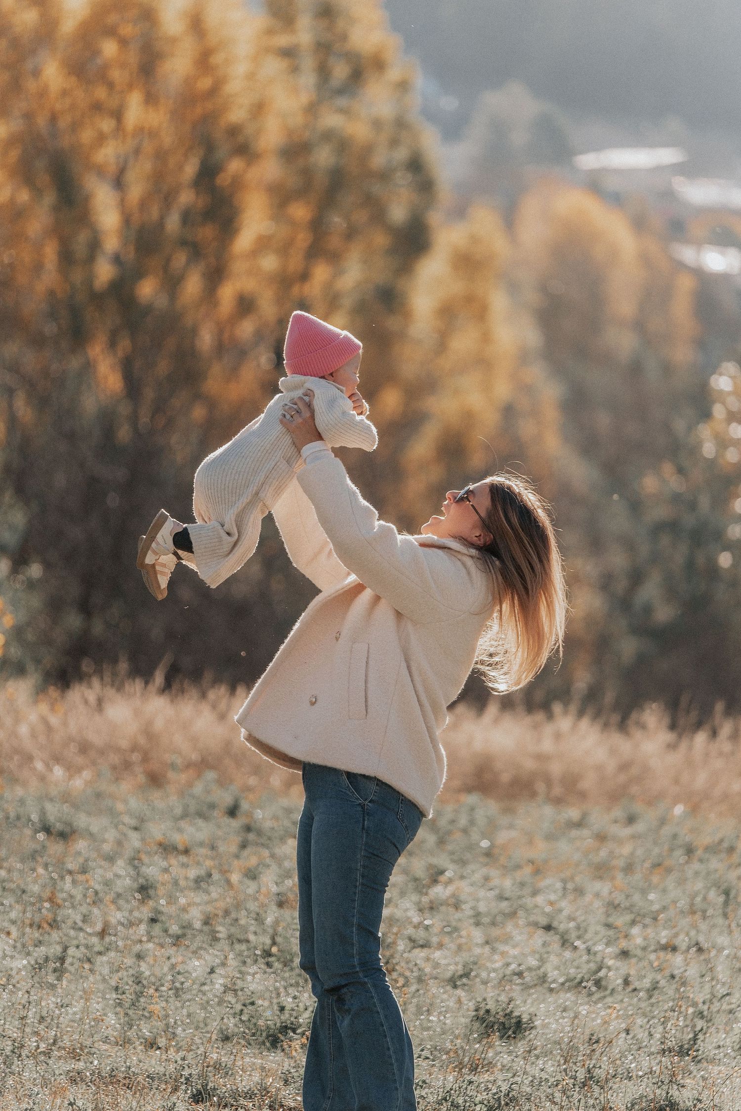 A person in a beige coat lifts a small baby wearing a pink hat against an autumn mountain backdrop at sunset.