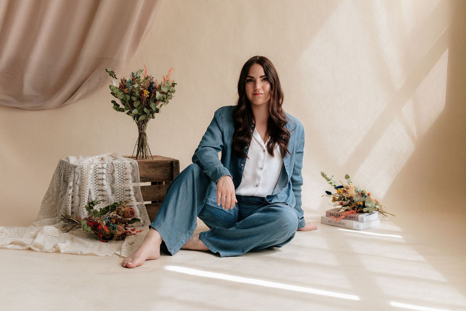 Woman in teal suit sitting cross-legged on floor with floral arrangements in a bright, airy studio setting.