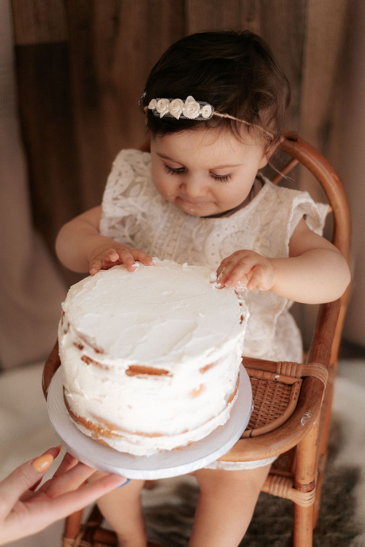 Baby in white lace dress reaches for a white frosted cake on a wooden chair in a warm indoor setting.