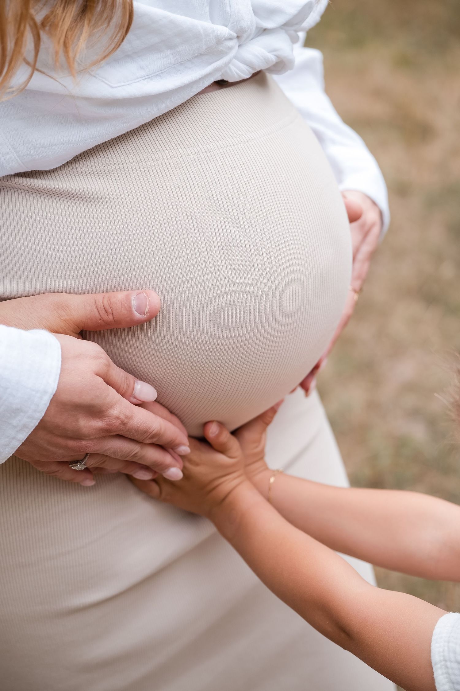A pregnant woman in a white sweater holds her belly while small hands reach to touch her baby bump.