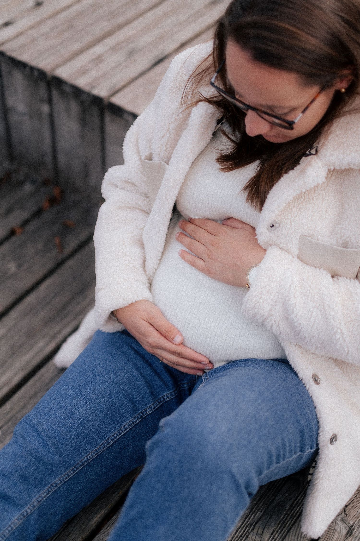 Pregnant woman in cozy white fleece coat and jeans sits on wooden deck while gently touching her baby bump.