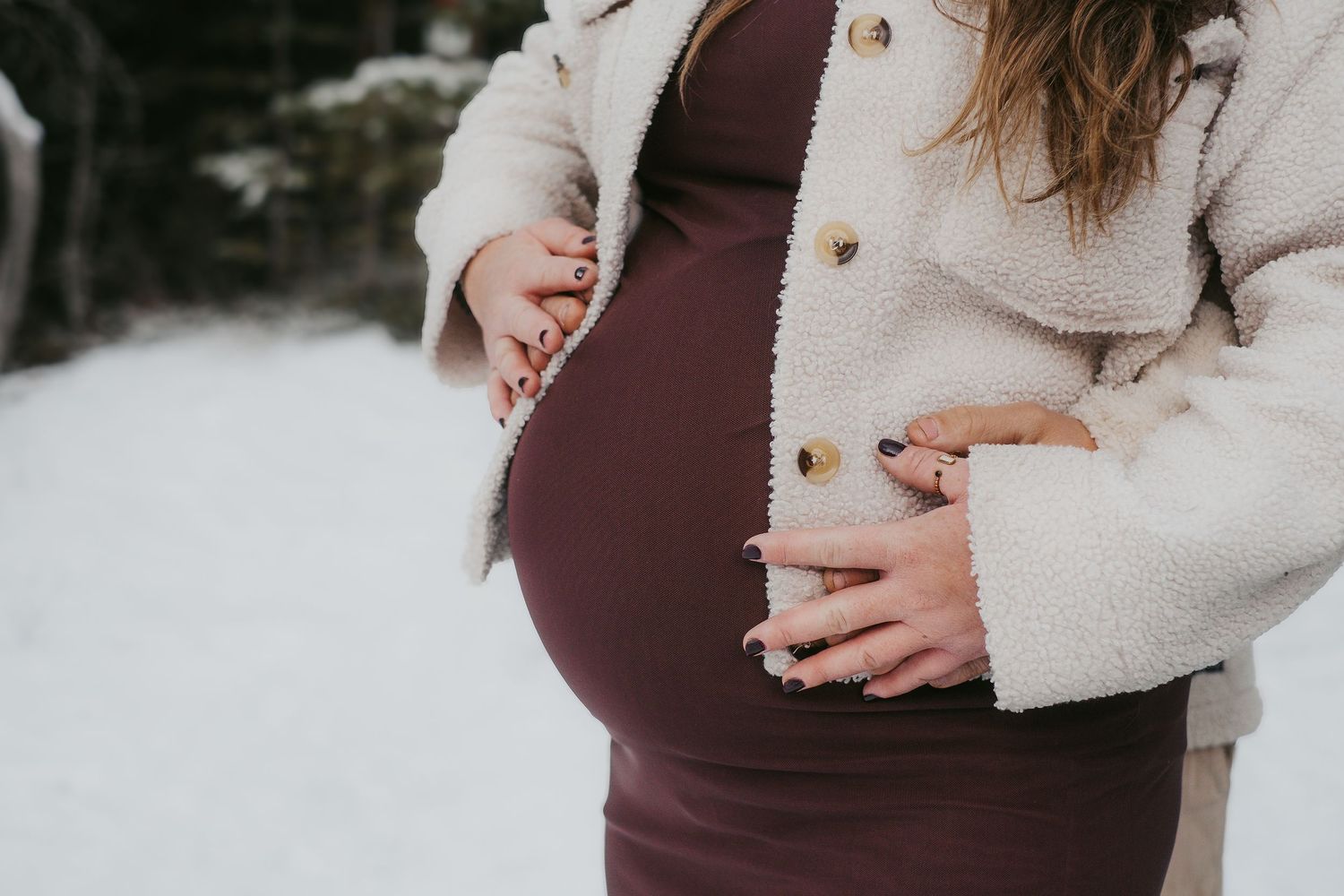 A pregnant person in a burgundy dress and cream teddy coat holds their belly in a snowy winter setting.