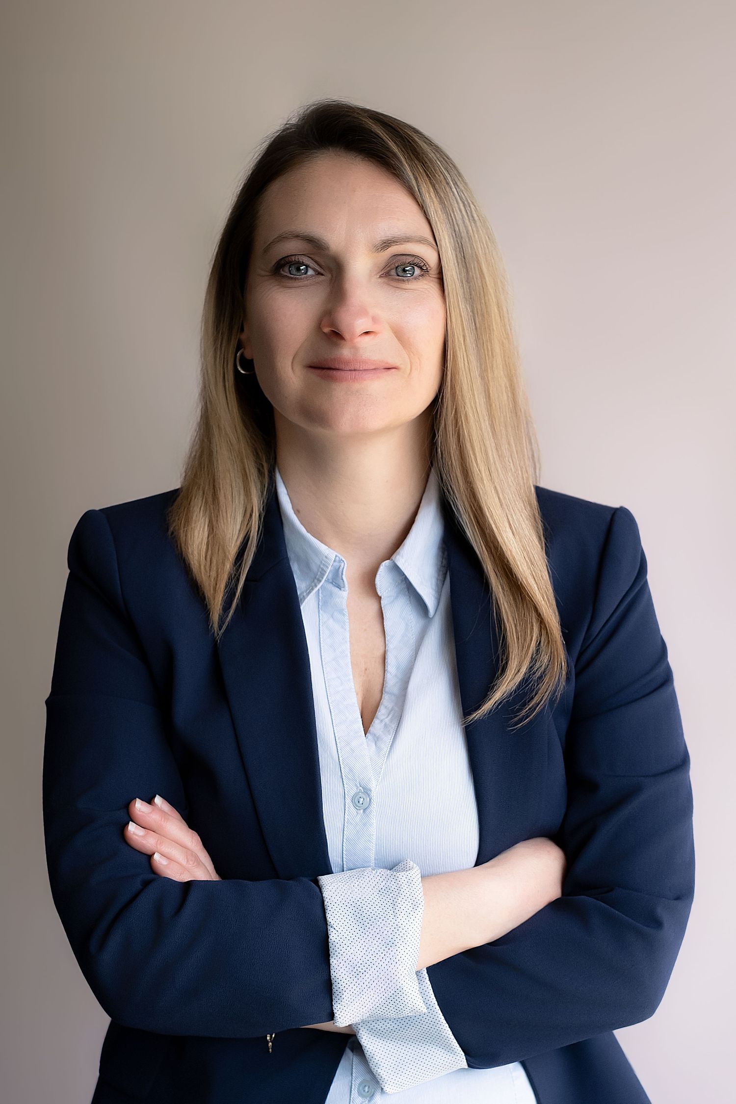 Professional woman in navy blazer and light blue shirt, arms crossed, smiling confidently against neutral background.