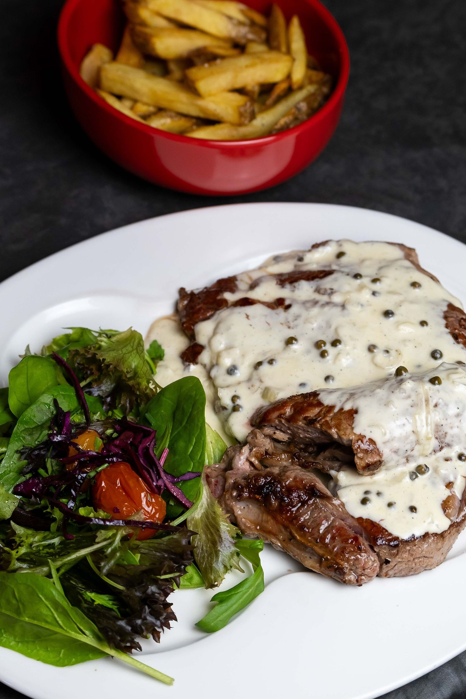 Grilled steak with creamy pepper sauce, fresh mixed salad, and golden french fries in a red bowl on a white plate.
