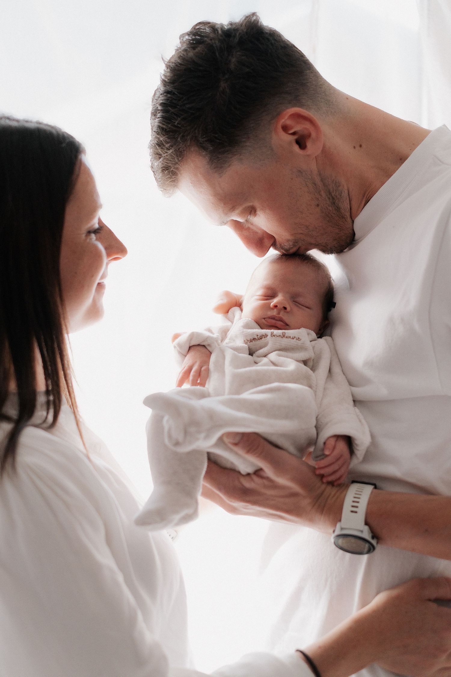 Parents in white clothing share an intimate moment with their newborn baby wrapped in a white blanket.