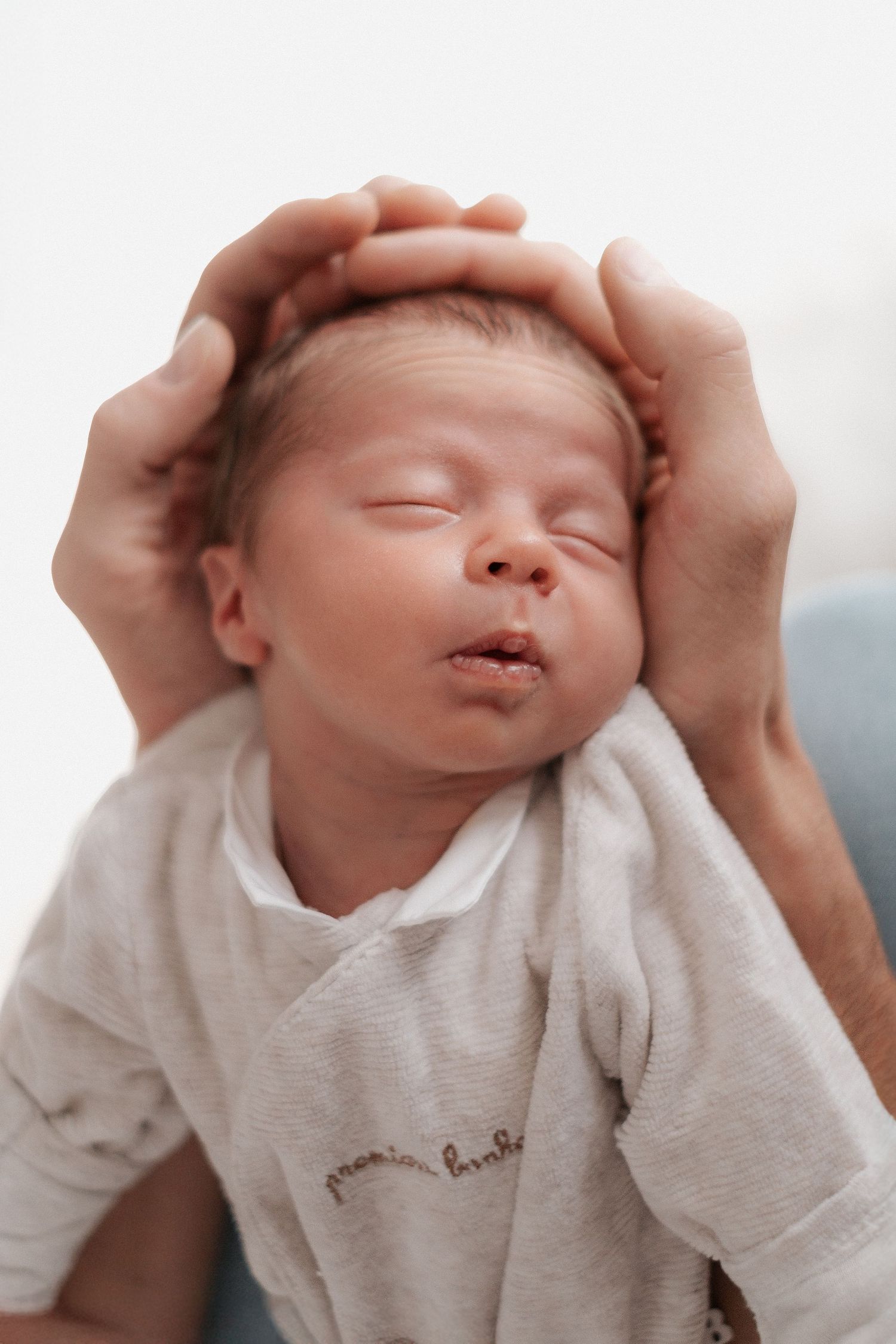 A sleeping newborn baby is gently cradled in caring hands wearing a soft white onesie.