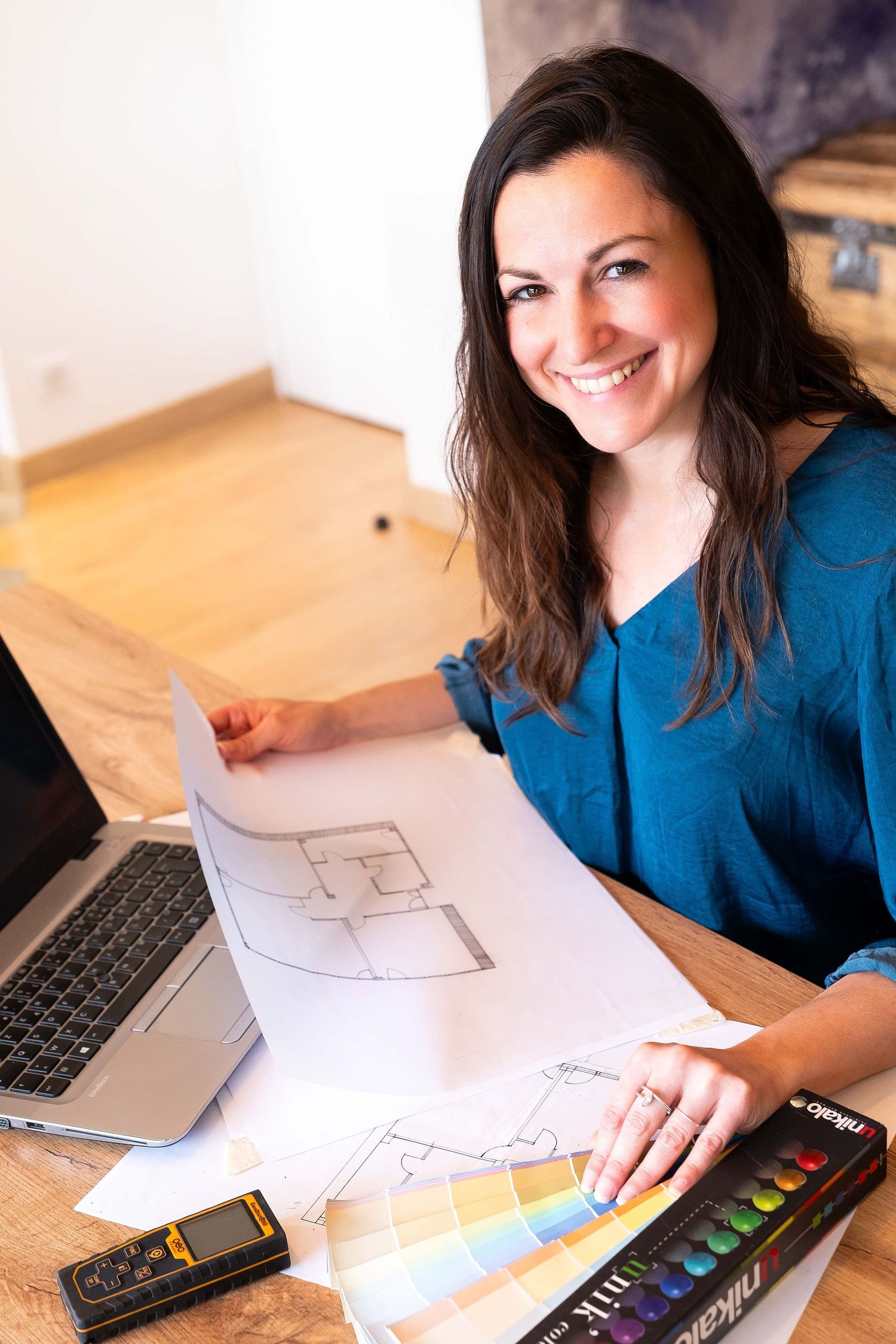 Smiling female interior designer reviewing floor plans with color swatches and laptop at wooden desk.
