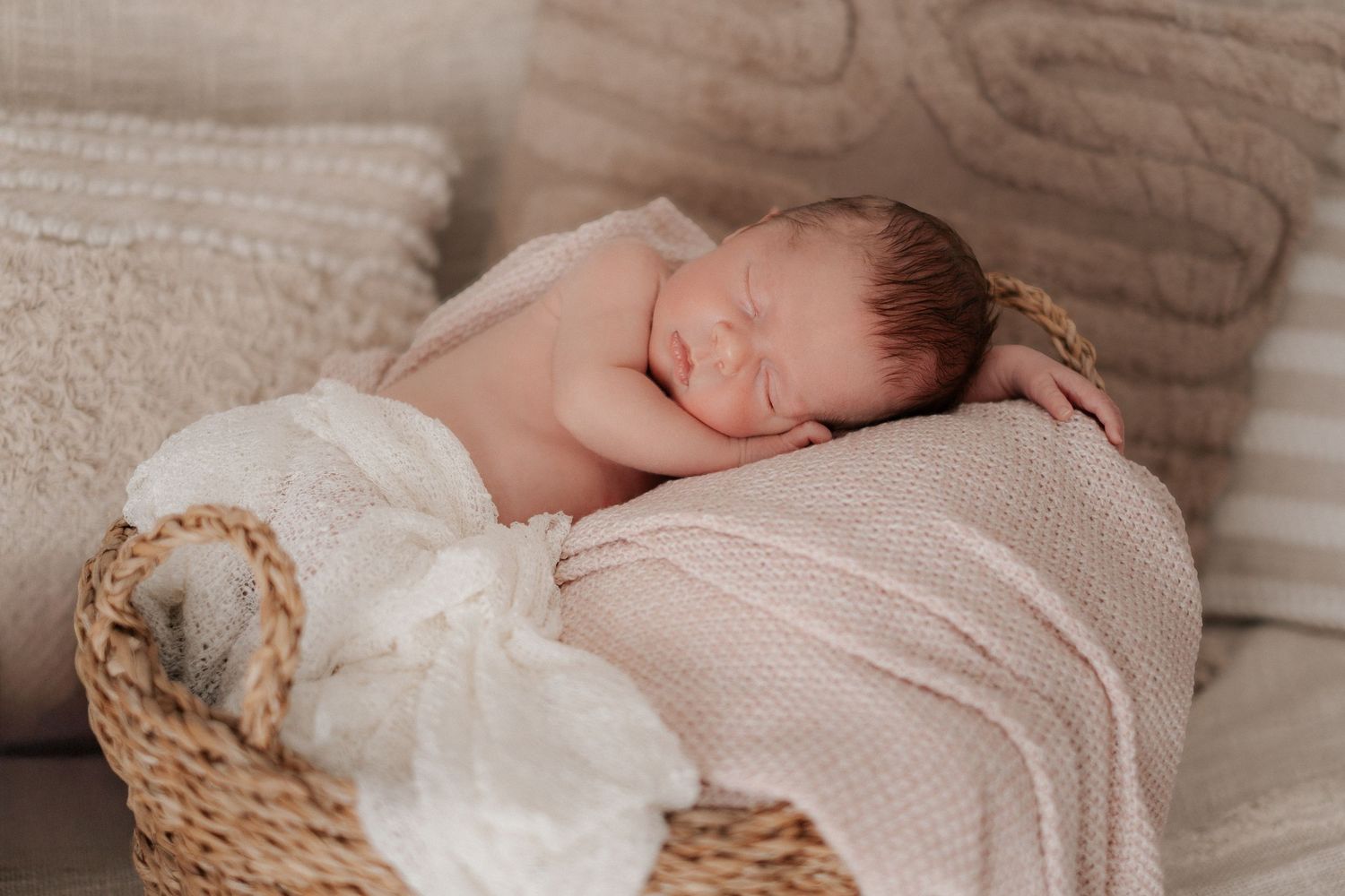 Newborn baby sleeping peacefully in woven basket with soft pink blanket and delicate lace wrap.