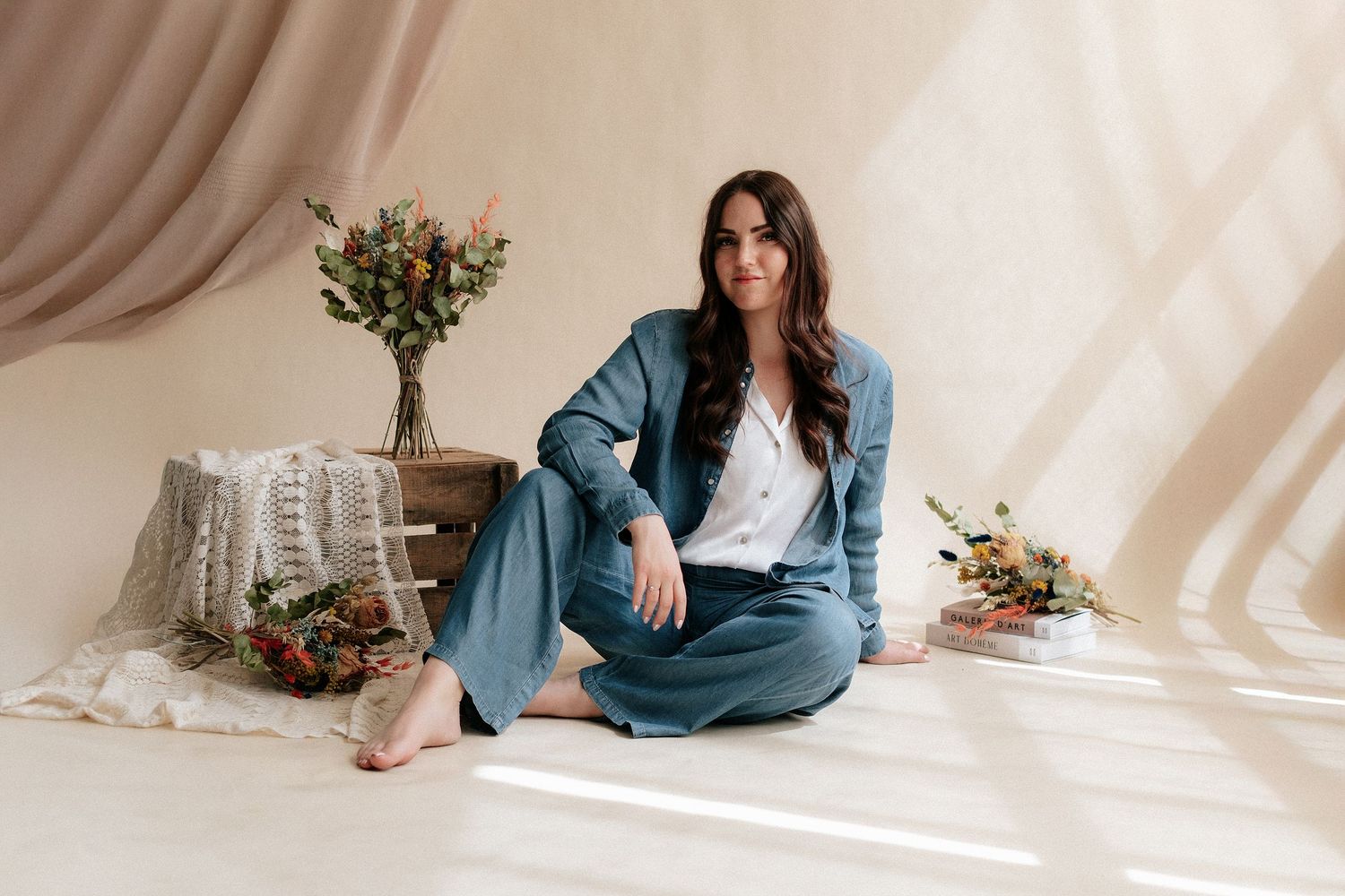 Woman in teal blazer and wide-leg pants sits cross-legged on floor with floral arrangements in bright studio.