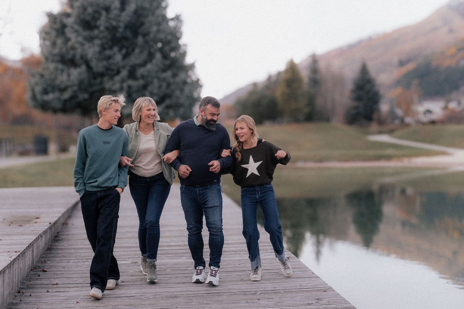 A group of friends joyfully runs along a lakeside path during autumn with mountains in the background.