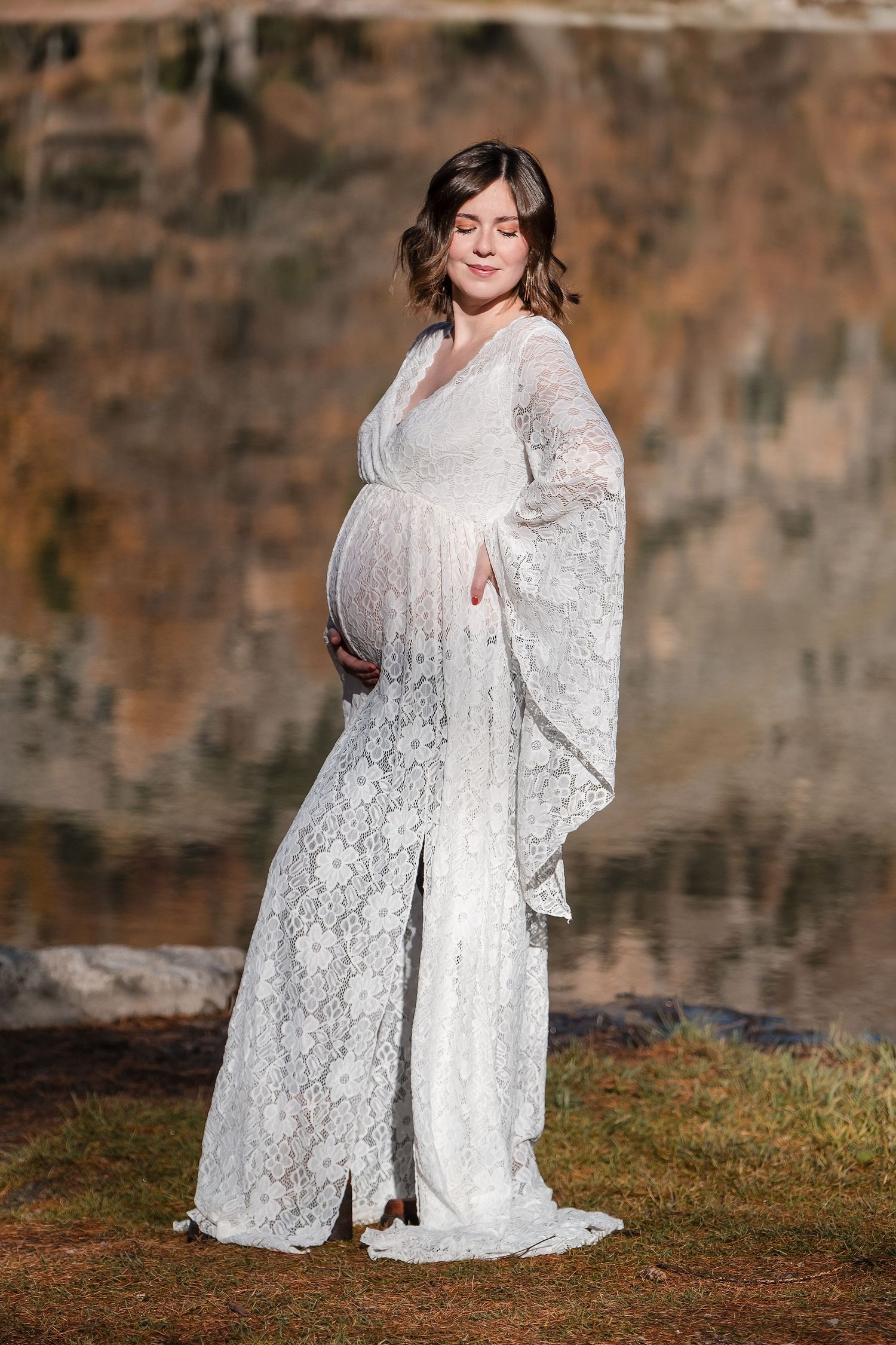 Expectant woman in flowing white lace maternity gown with bell sleeves poses outdoors against rocky backdrop.