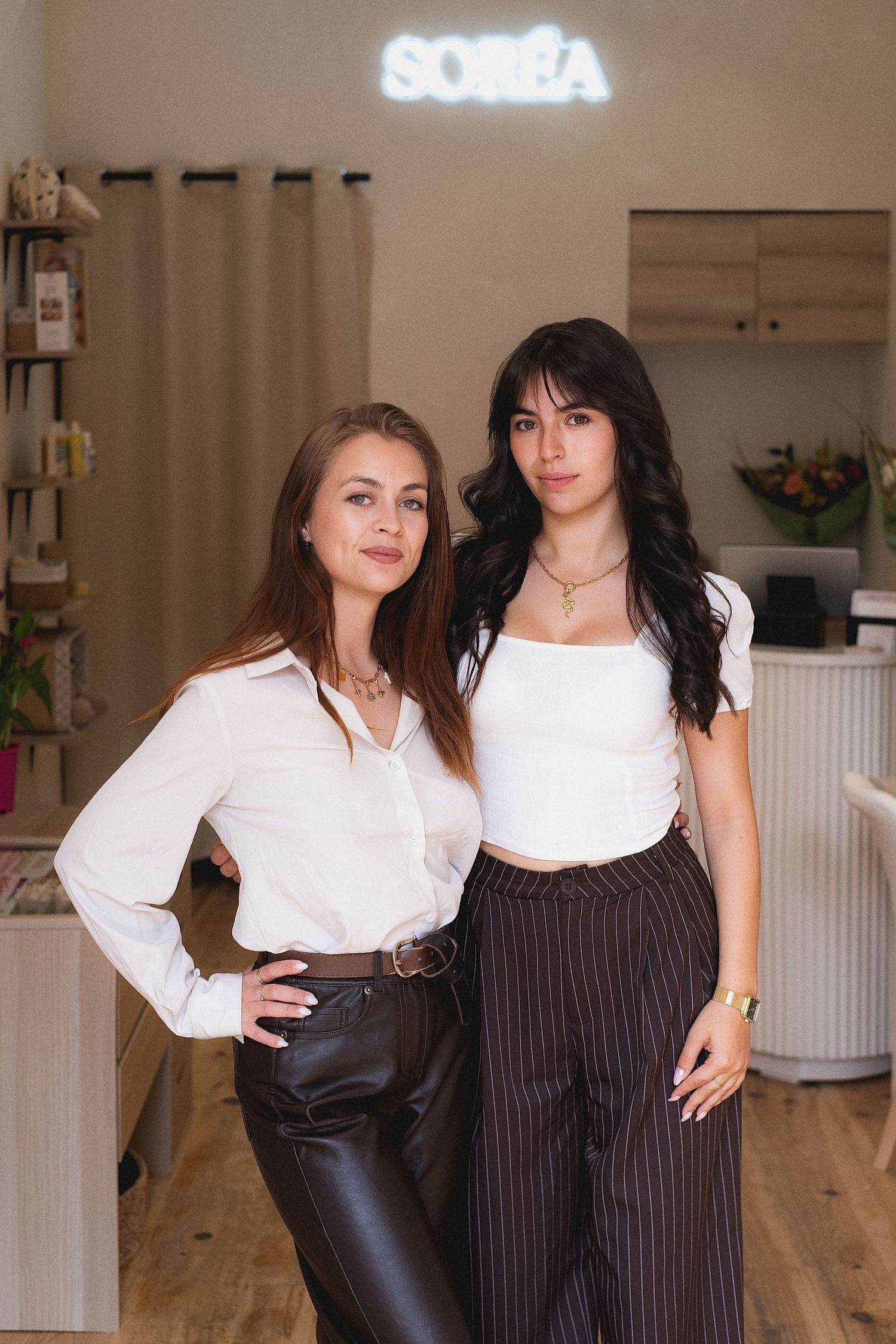 Two women in white tops pose together inside the Soréa beauty salon, one in leather pants and one in striped trousers.