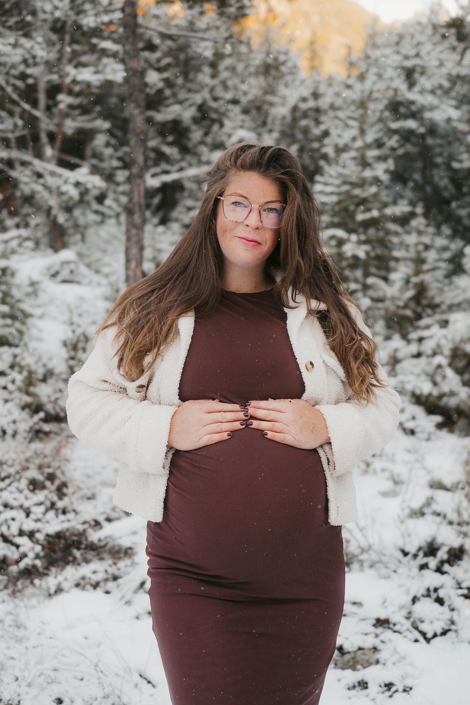 A person in a brown dress and white cardigan poses in a snowy forest setting during winter.
