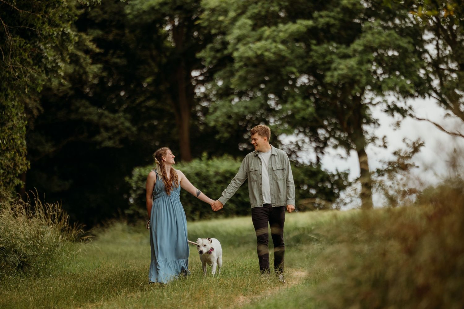 A couple walking their small white dog on a nature path while holding hands.