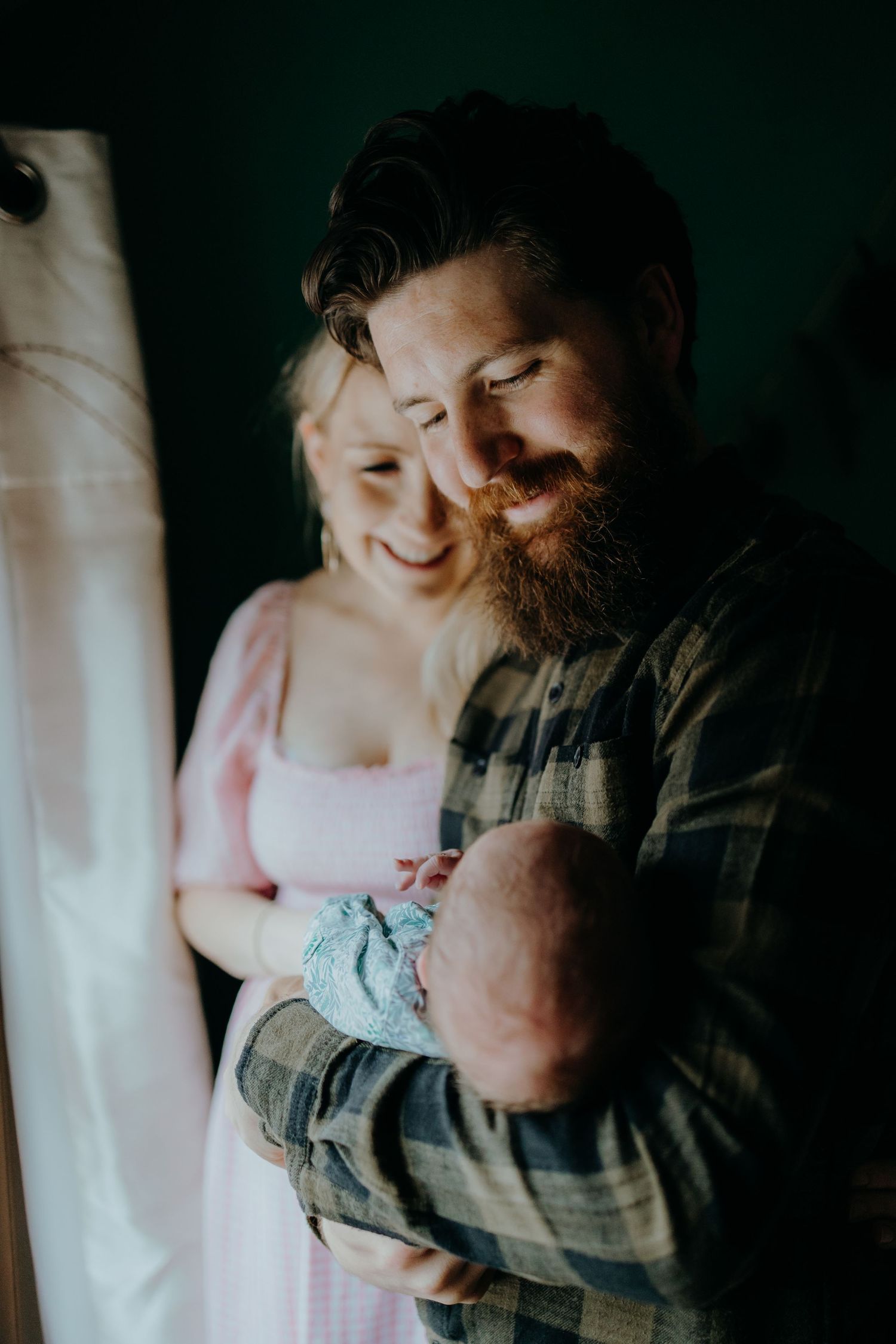 A tender family moment captured in moody lighting by a window as parents hold their newborn baby.