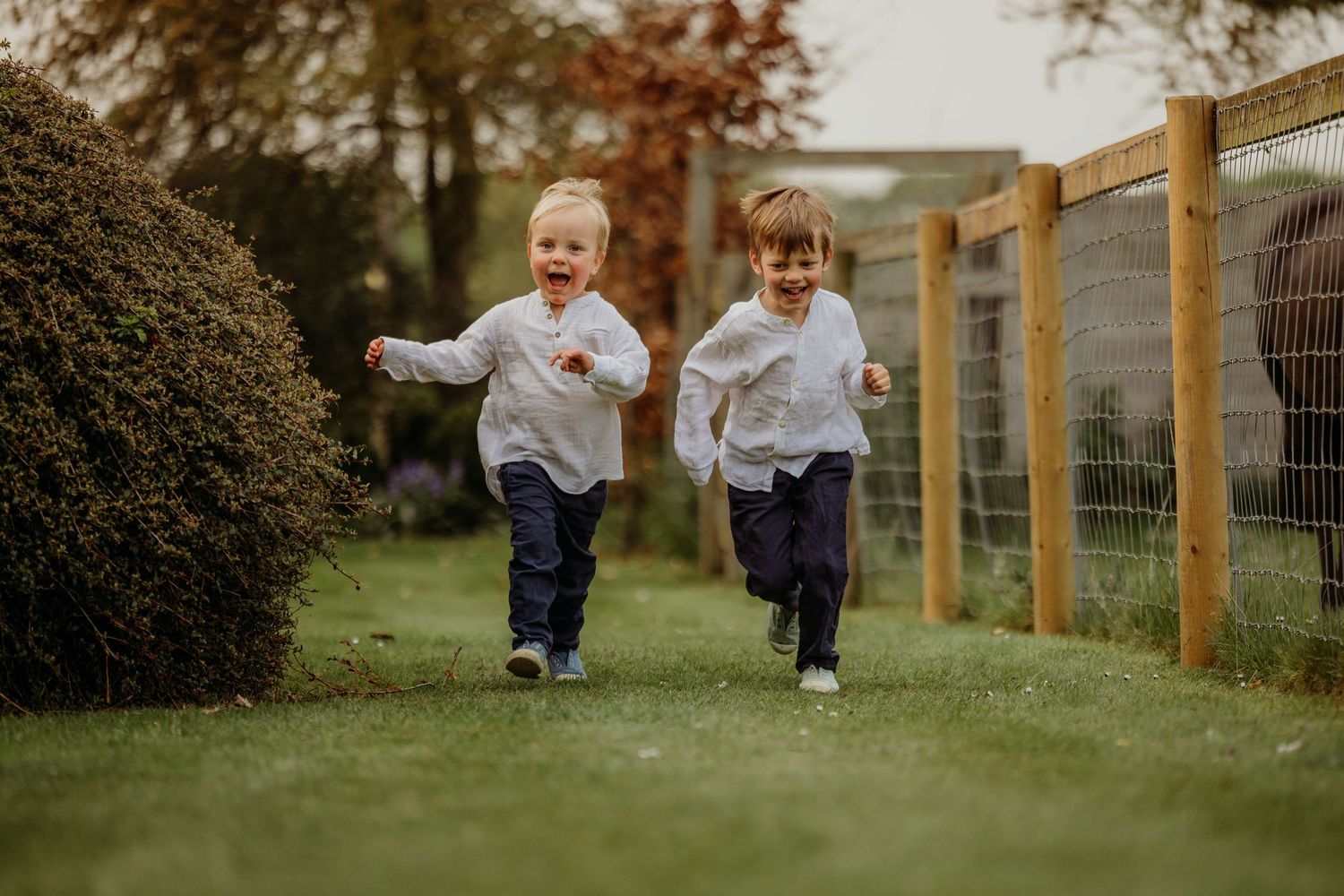 Two children running and laughing together along a grassy path with a wooden fence.