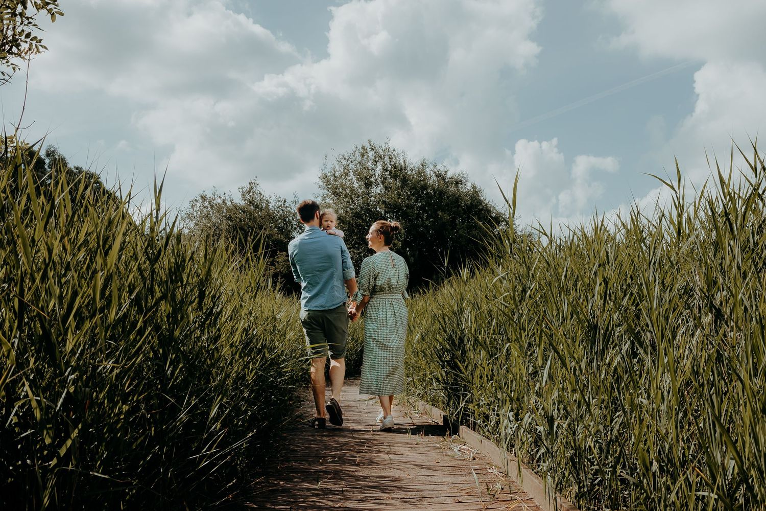 Two people walk hand in hand down a nature path surrounded by tall grass and greenery on a cloudy summer day.