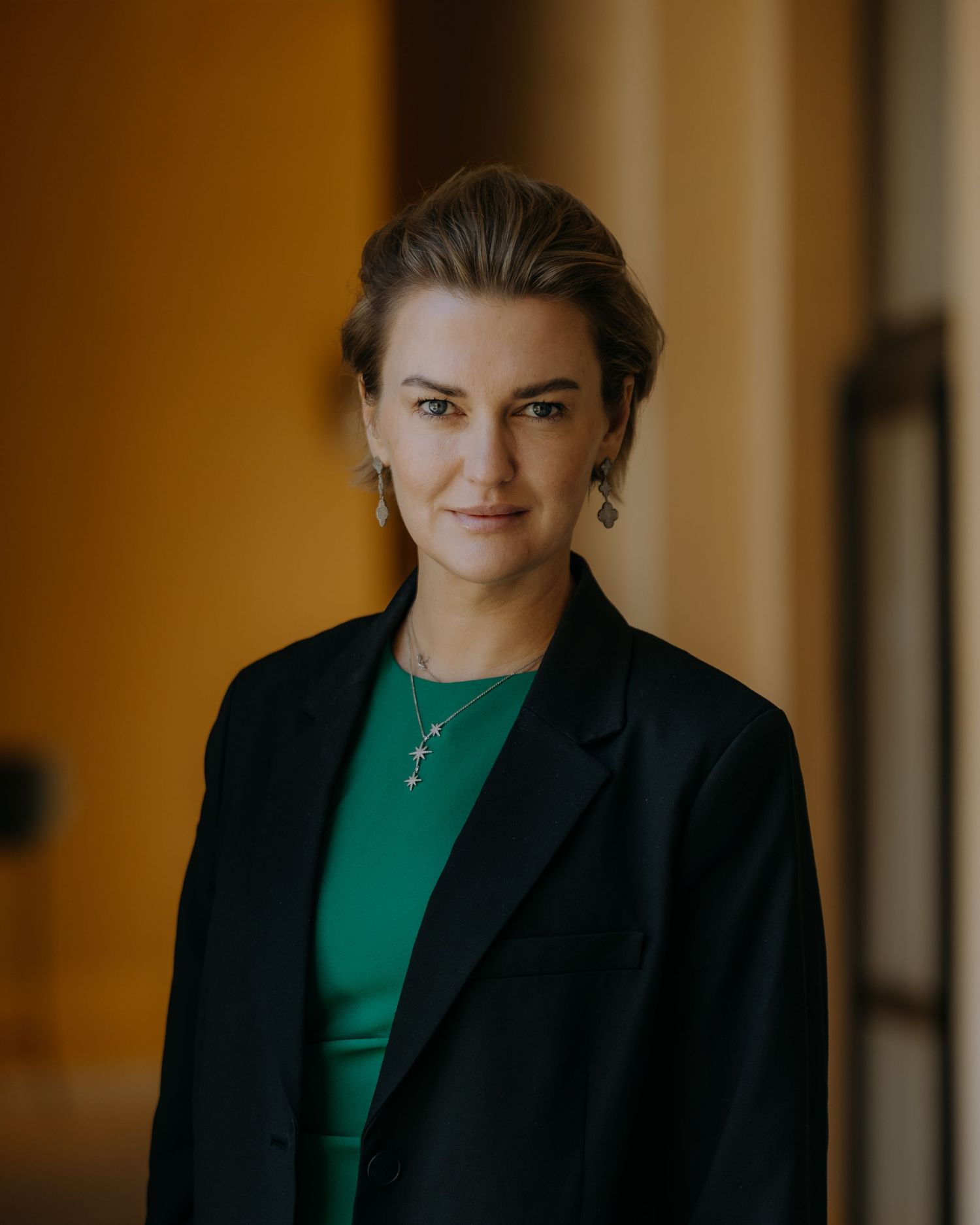 Professional portrait of person in green top and black blazer against warm-toned background.
