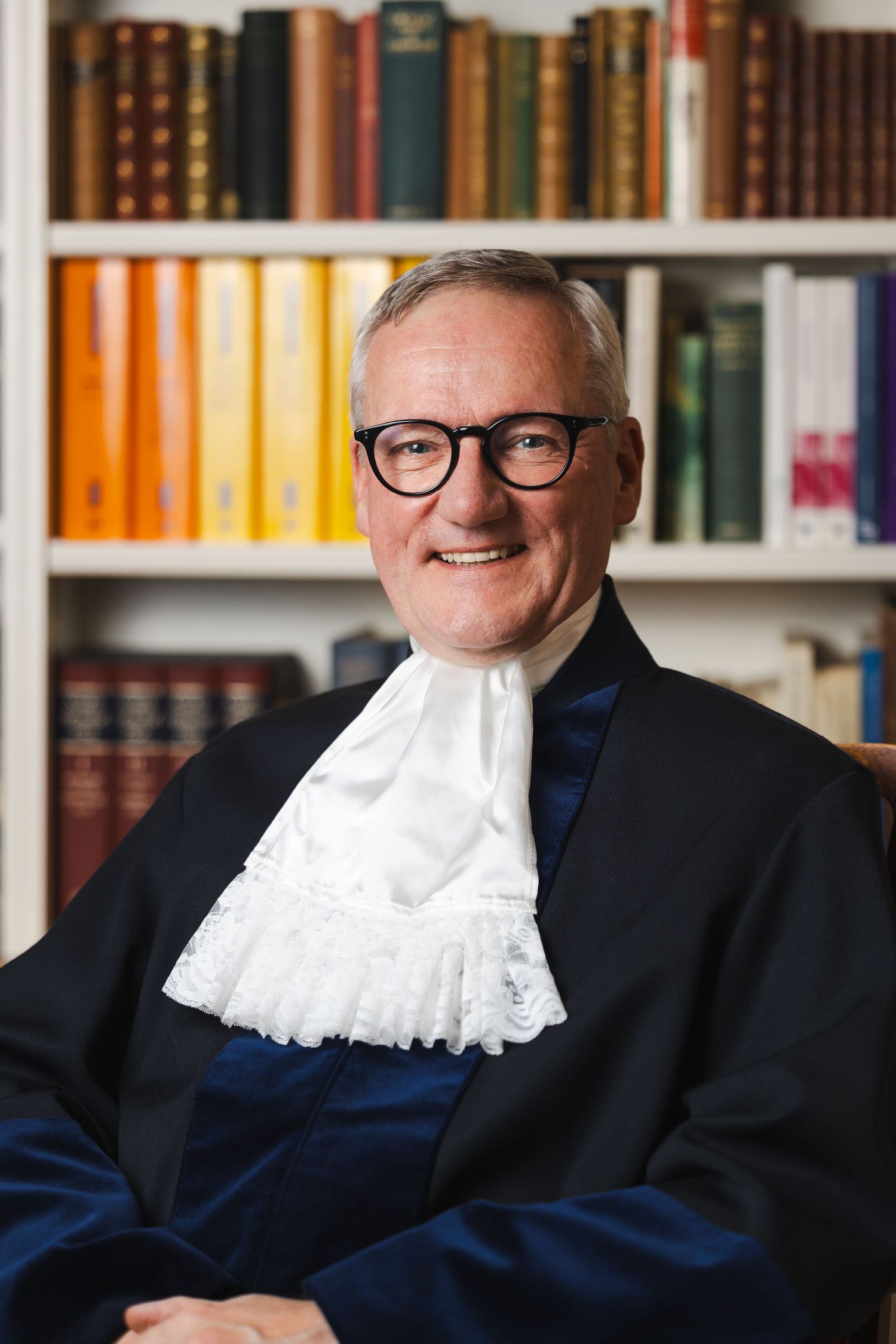 Legal professional wearing traditional court dress with jabot and glasses in front of library.