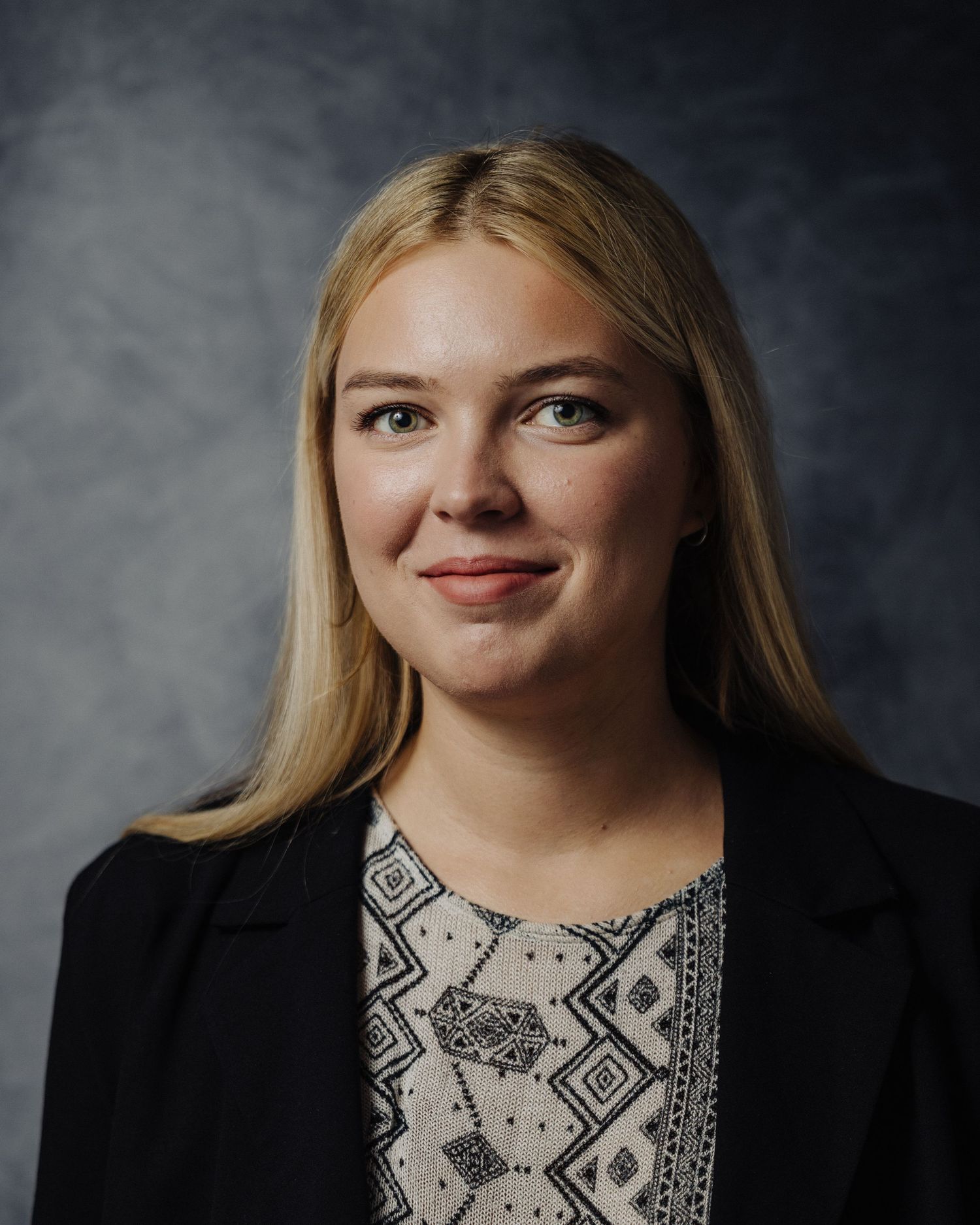 Studio portrait with person wearing black blazer and patterned top against dark background.