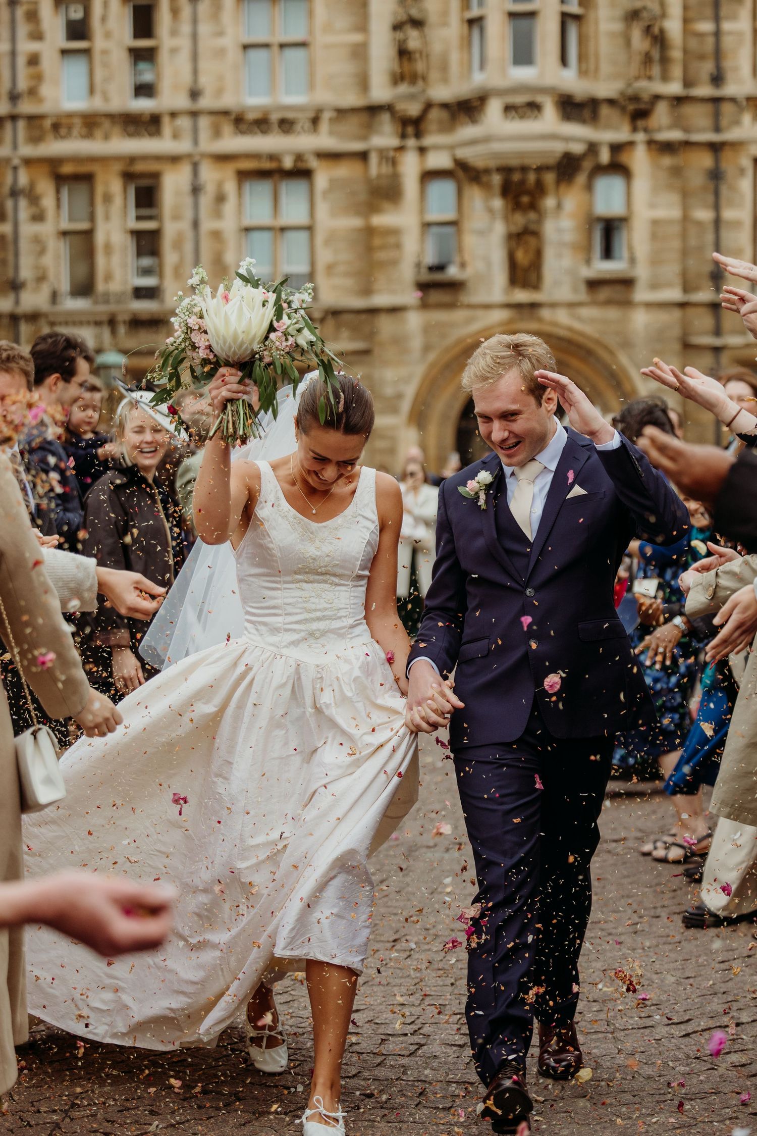 Wedding party running through confetti shower on cobblestone path outside historic building.