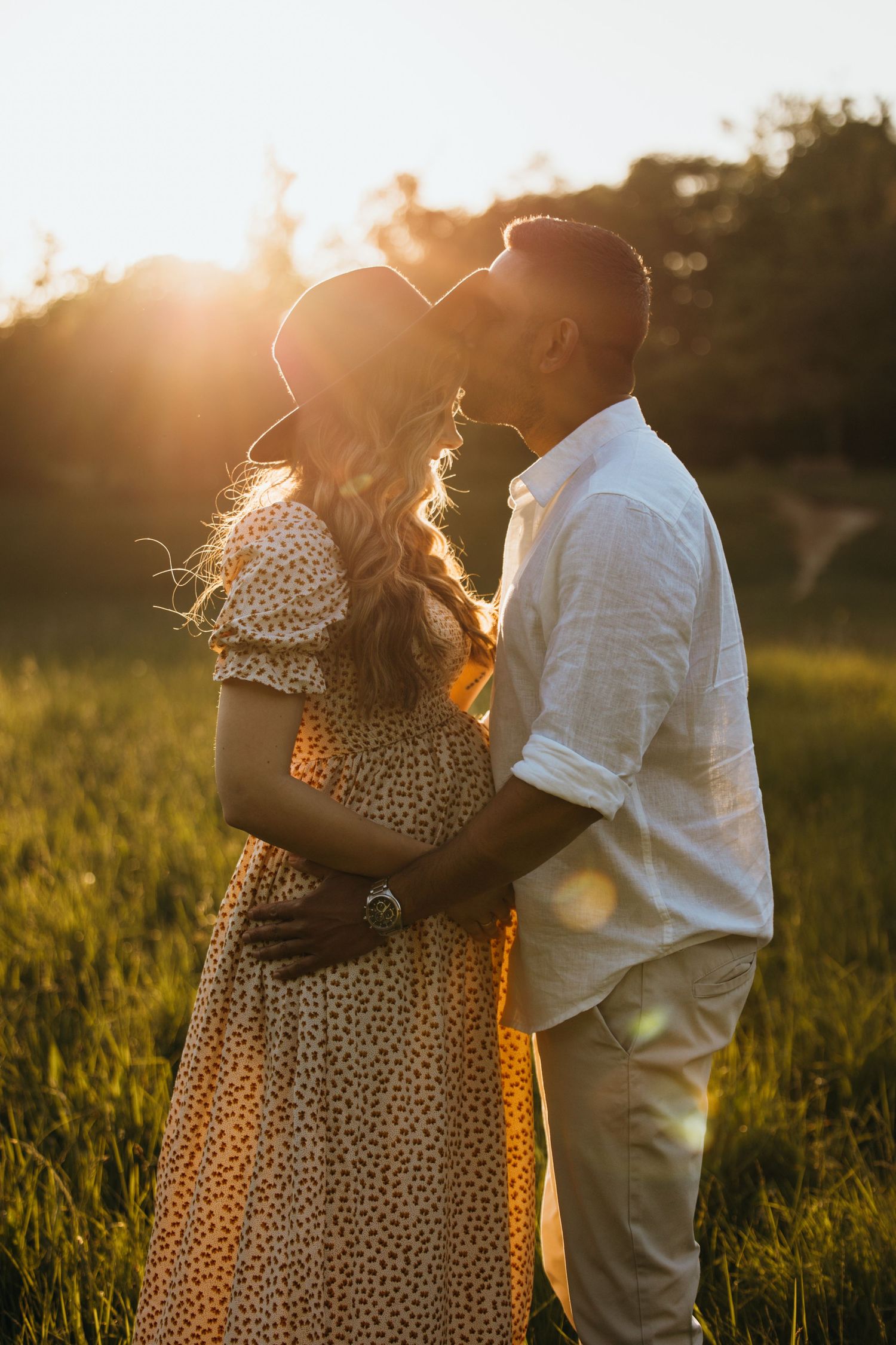 Couple embraces in golden sunlit meadow wearing polka dot dress and white shirt at sunset.