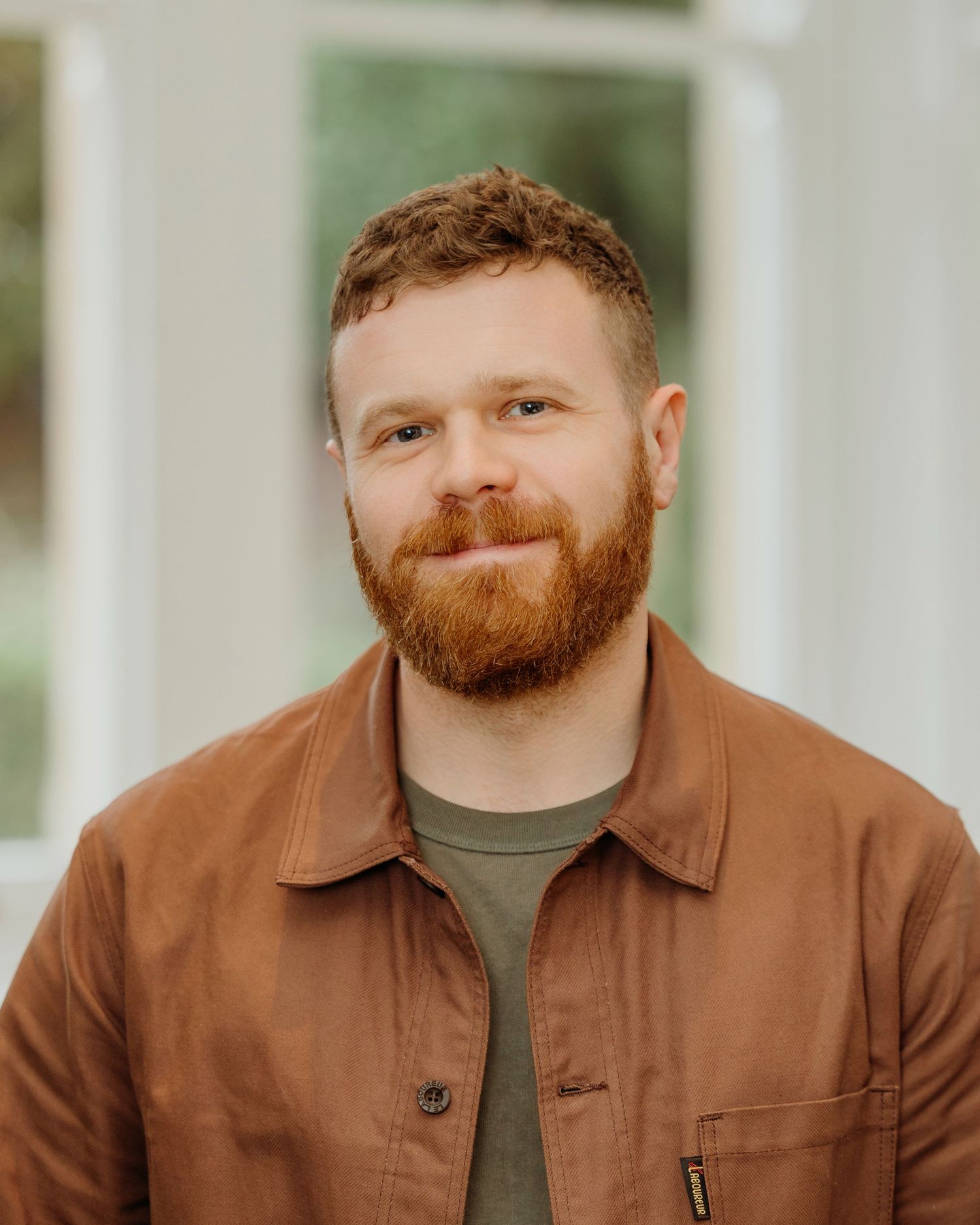 Portrait of person in brown jacket and dark shirt against bright indoor background.