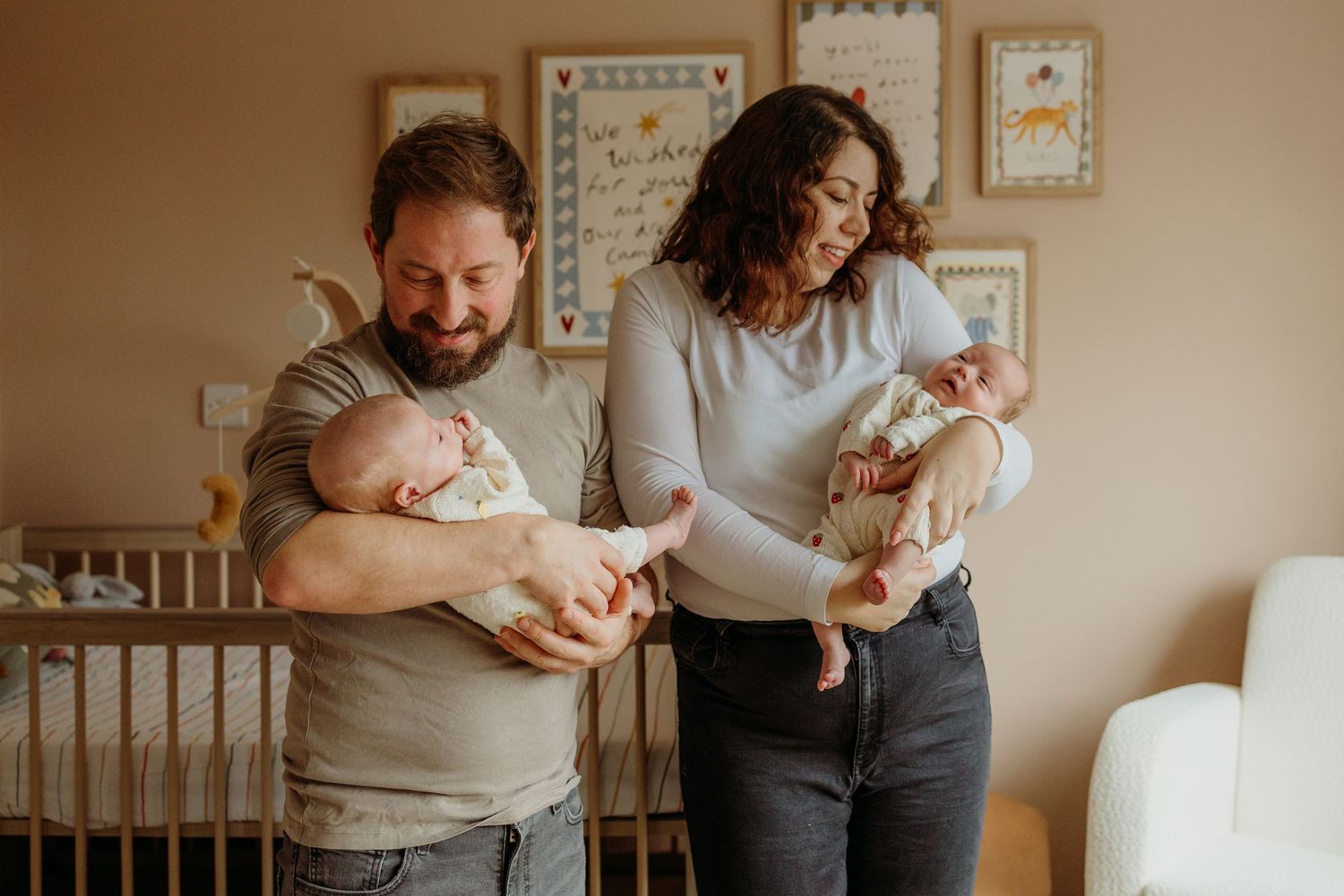 Two adults hold and comfort their newborn twins in a cozy nursery with framed artwork on the wall.
